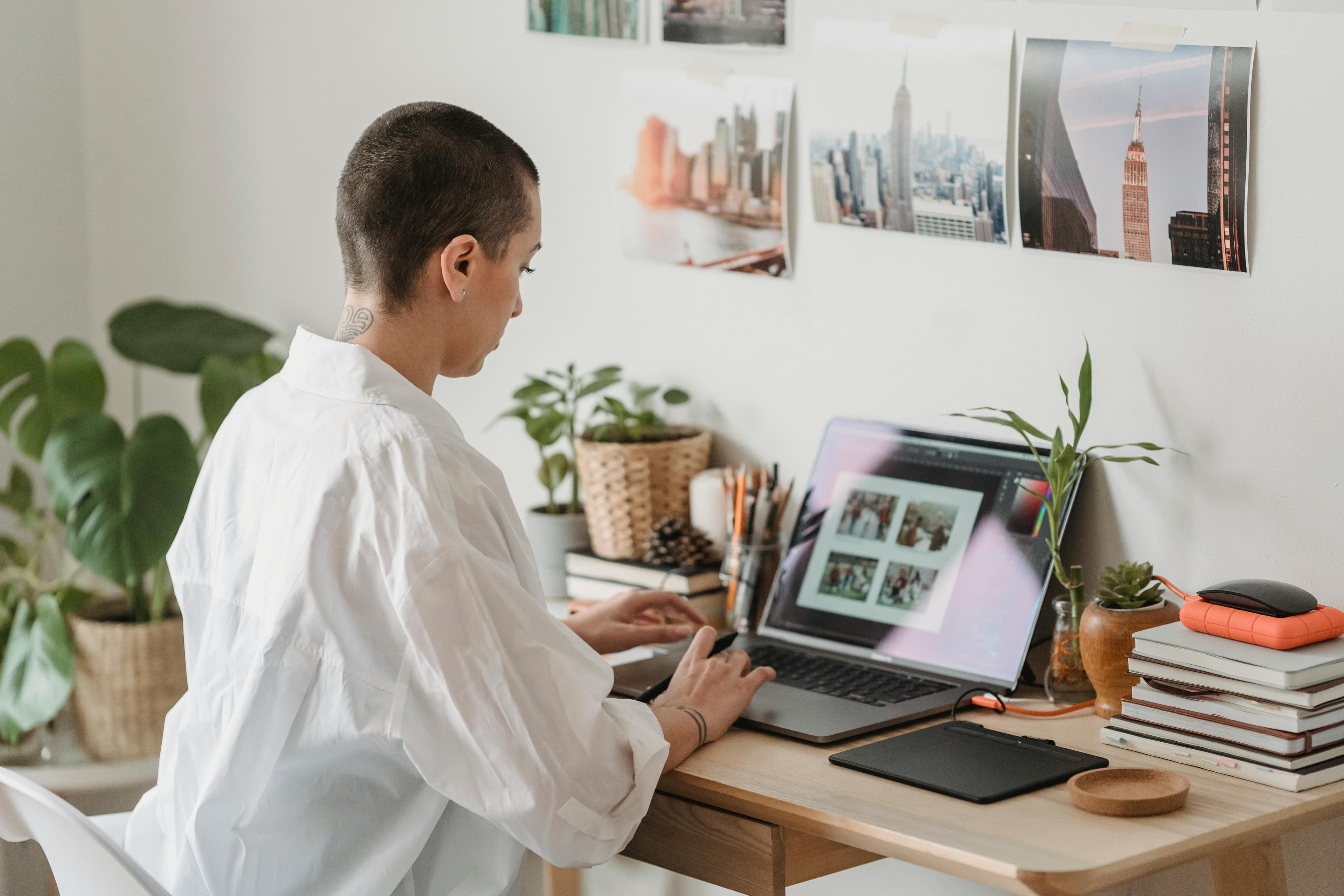 A person with short hair, dressed in a white shirt, working on a laptop at a desk decorated with plants and cityscape photos. They appear focused on designing marketing materials, showcasing the creative work environment of a marketing and promotion agency.