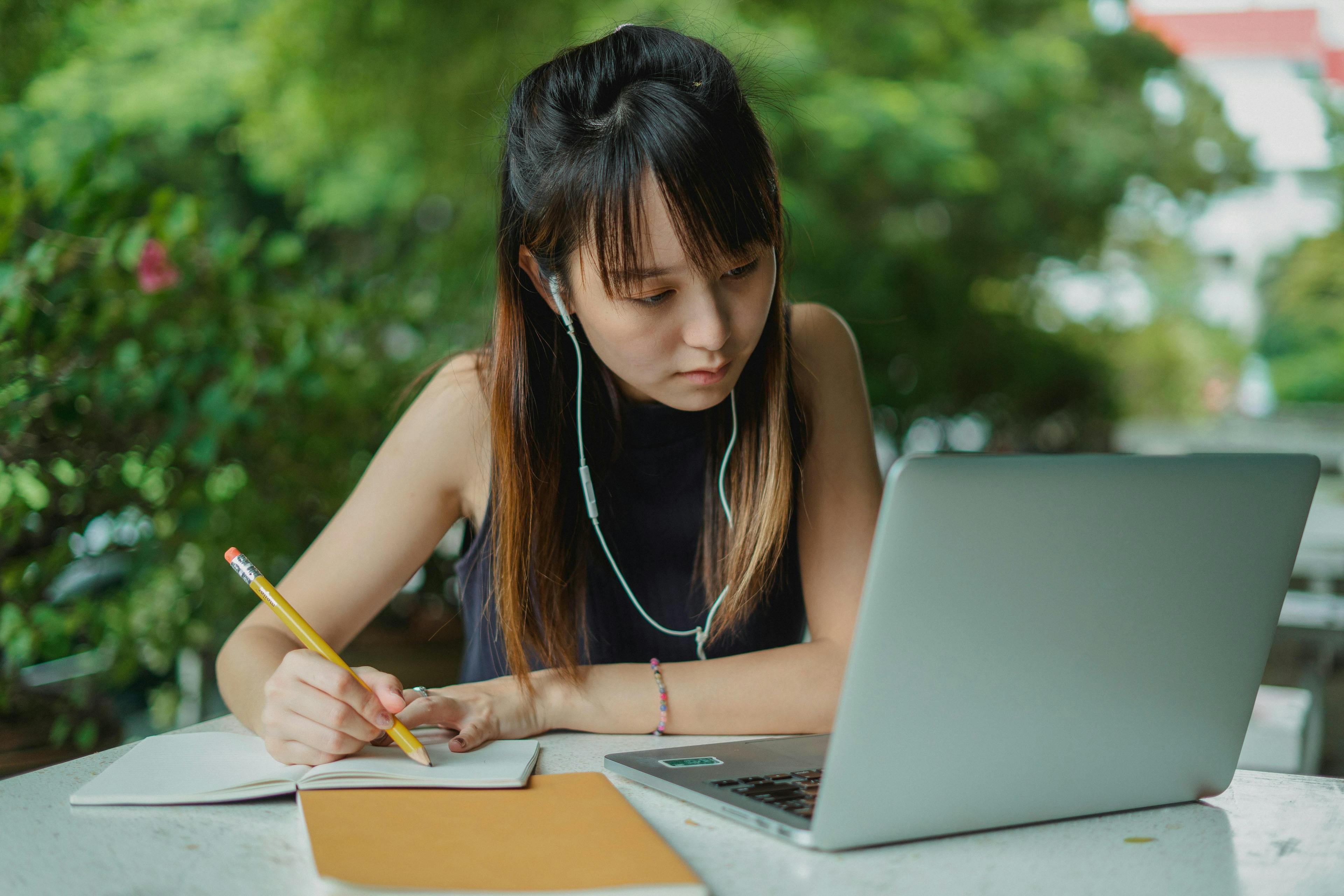 A young woman working on her laptop while taking notes in a notebook, set in a peaceful outdoor environment. This image is perfect for illustrating the "leader VS manager" concept, emphasizing a leader's ability to work efficiently in various settings.