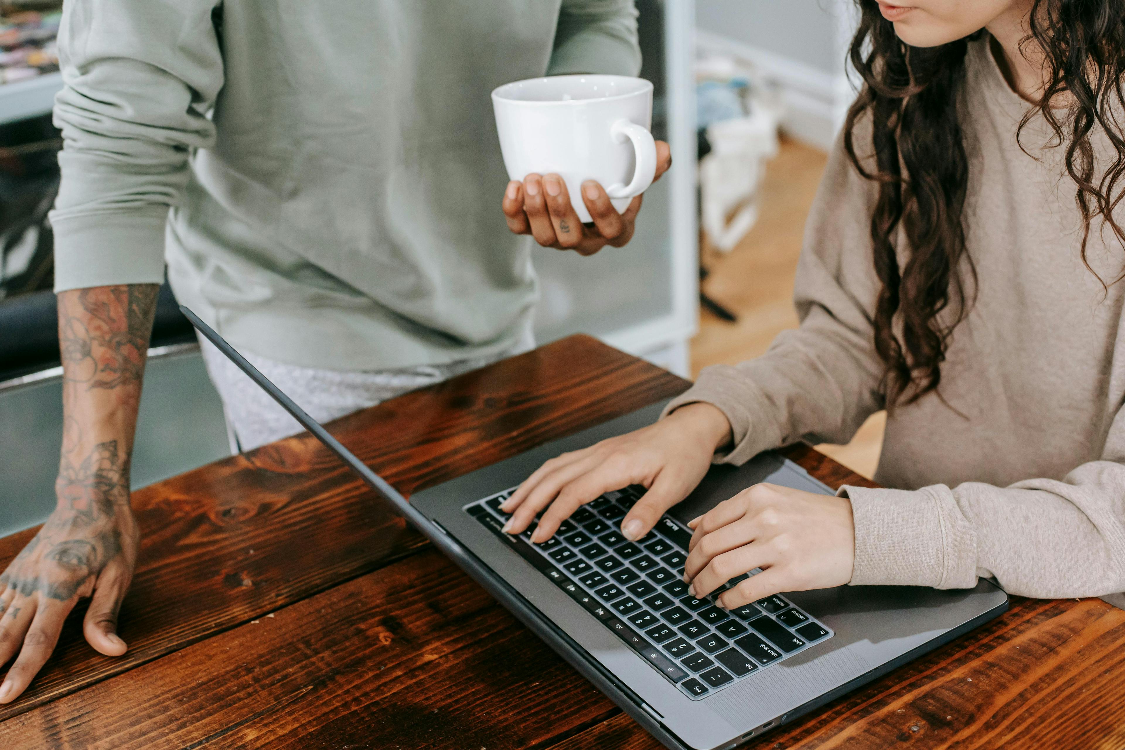 Two individuals collaborating at a wooden table, one holding a coffee cup while the other types on a laptop. This image is ideal for lead magnetic materials emphasizing teamwork and collaborative efforts in a relaxed setting.