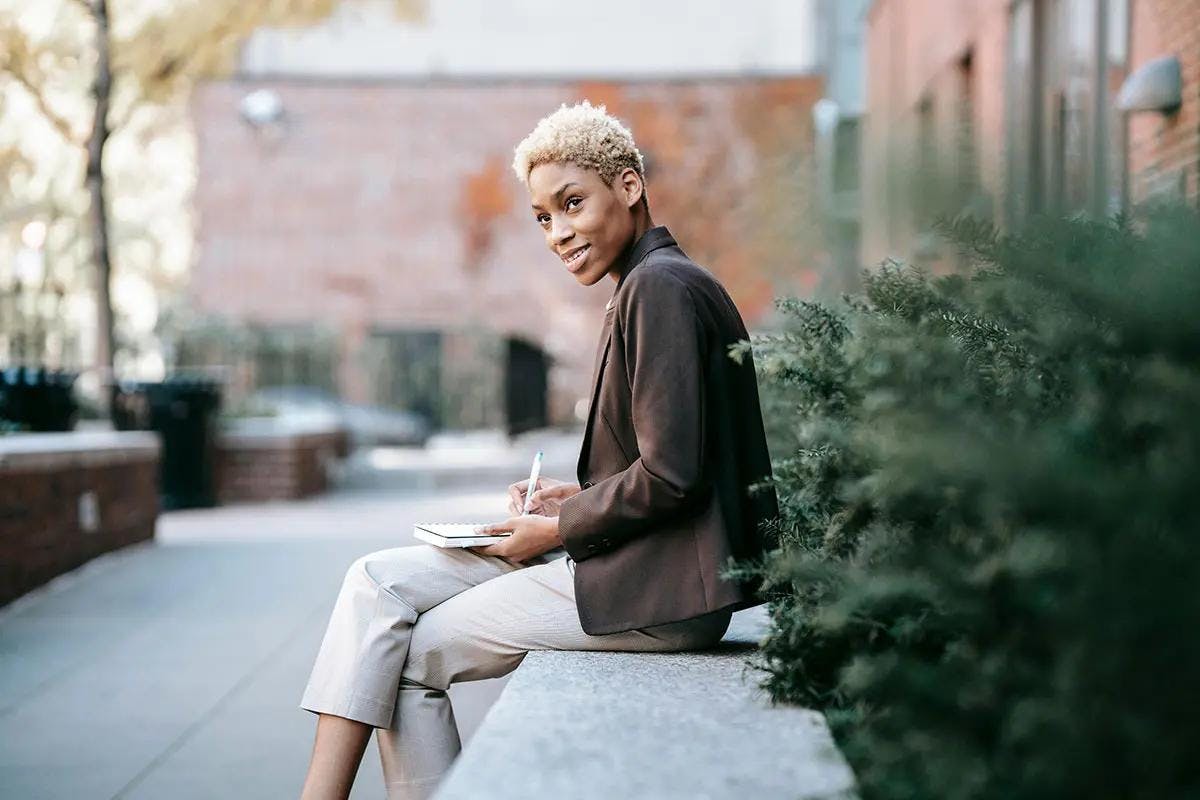 A confident professional sits on an outdoor bench, jotting down notes in a notebook. She has short, blonde hair and is dressed in a smart brown blazer with light-colored trousers. The background features urban greenery and buildings, creating a serene yet professional atmosphere. This image is ideal for conveying the concept of "B2B SaaS marketing."
