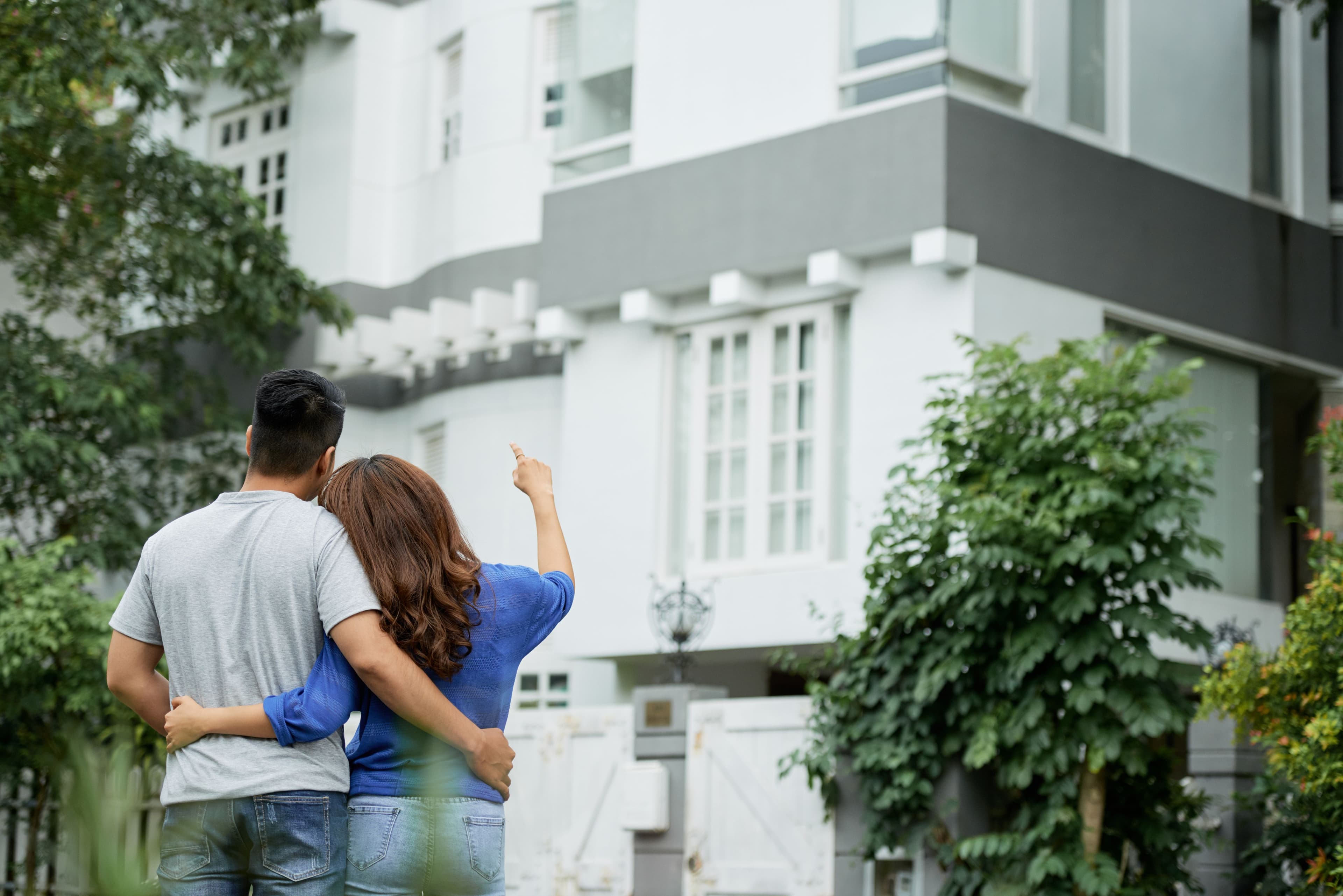 A young couple stands arm in arm, gazing at their dream house behind a gated fence, considering the purchase. This illustrates split testing for pricing by comparing different property values or investment options.