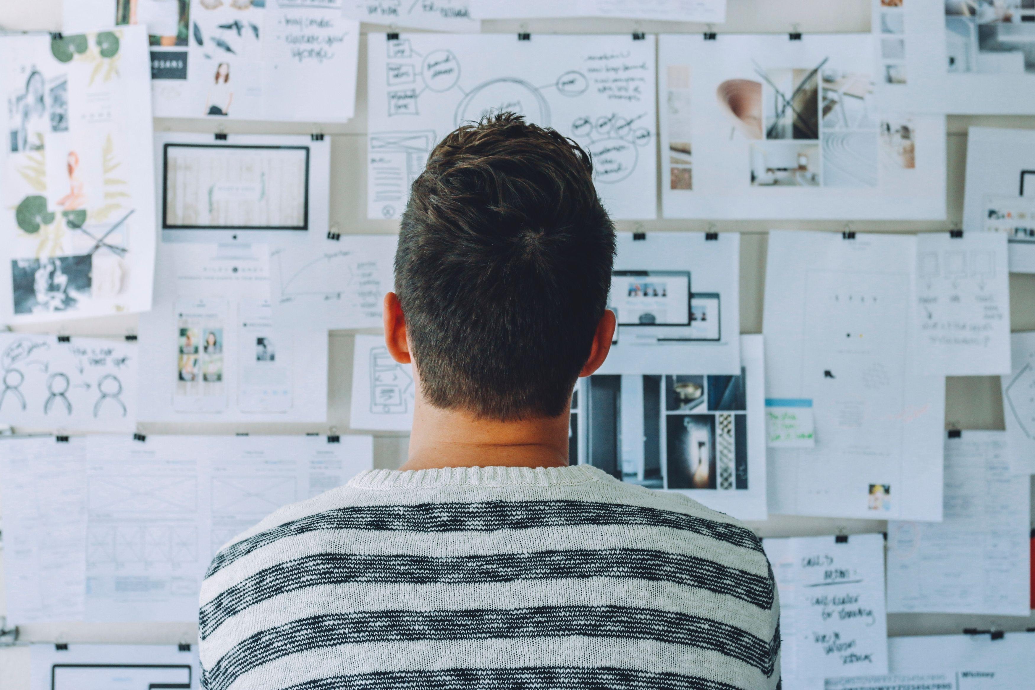 A man with short hair facing a wall filled with notes, sketches, and diagrams pinned up for brainstorming or project planning. He seems to be analyzing the content for insights.