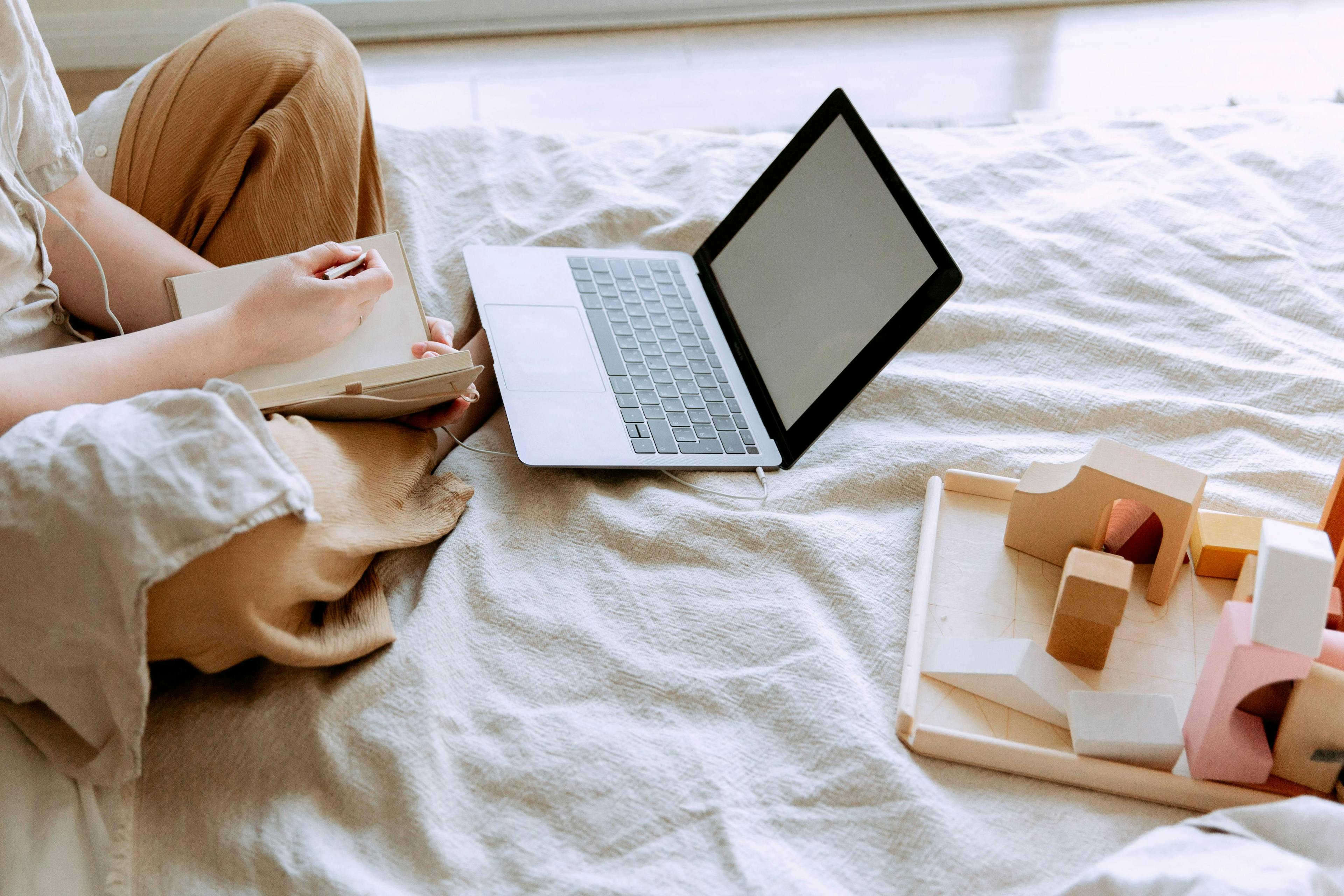 a person sitting on a bed with a laptop, writing in a notebook, and next to them are wooden building blocks. The person seems to be working from home, and a question arises: "Is lead magnetic?"