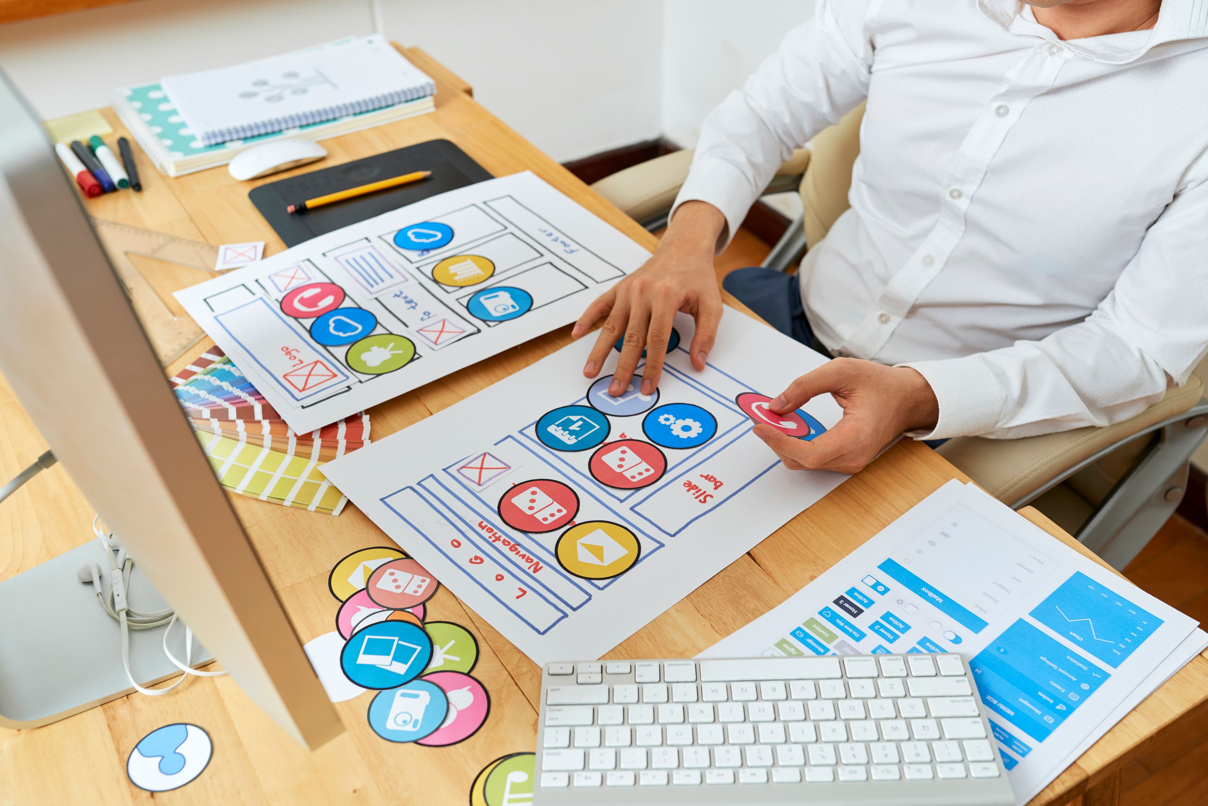  A designer works at a desk, arranging colorful icons and app interface layouts. This scene represents startup marketing through developing an engaging and user-friendly mobile app experience to attract and retain customers.