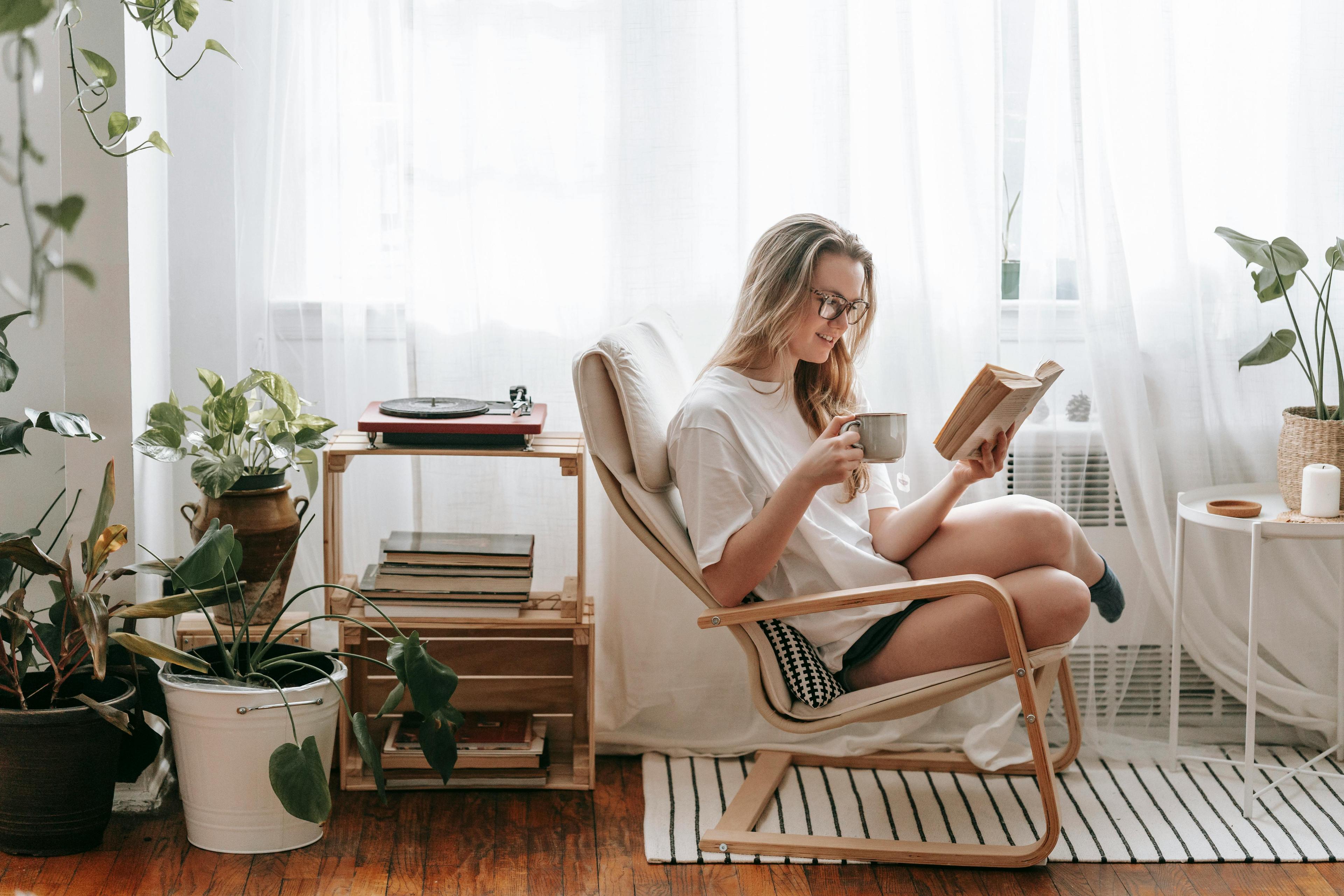 A woman enjoying a peaceful moment, reading a book and drinking coffee in a bright, cozy room filled with indoor plants.