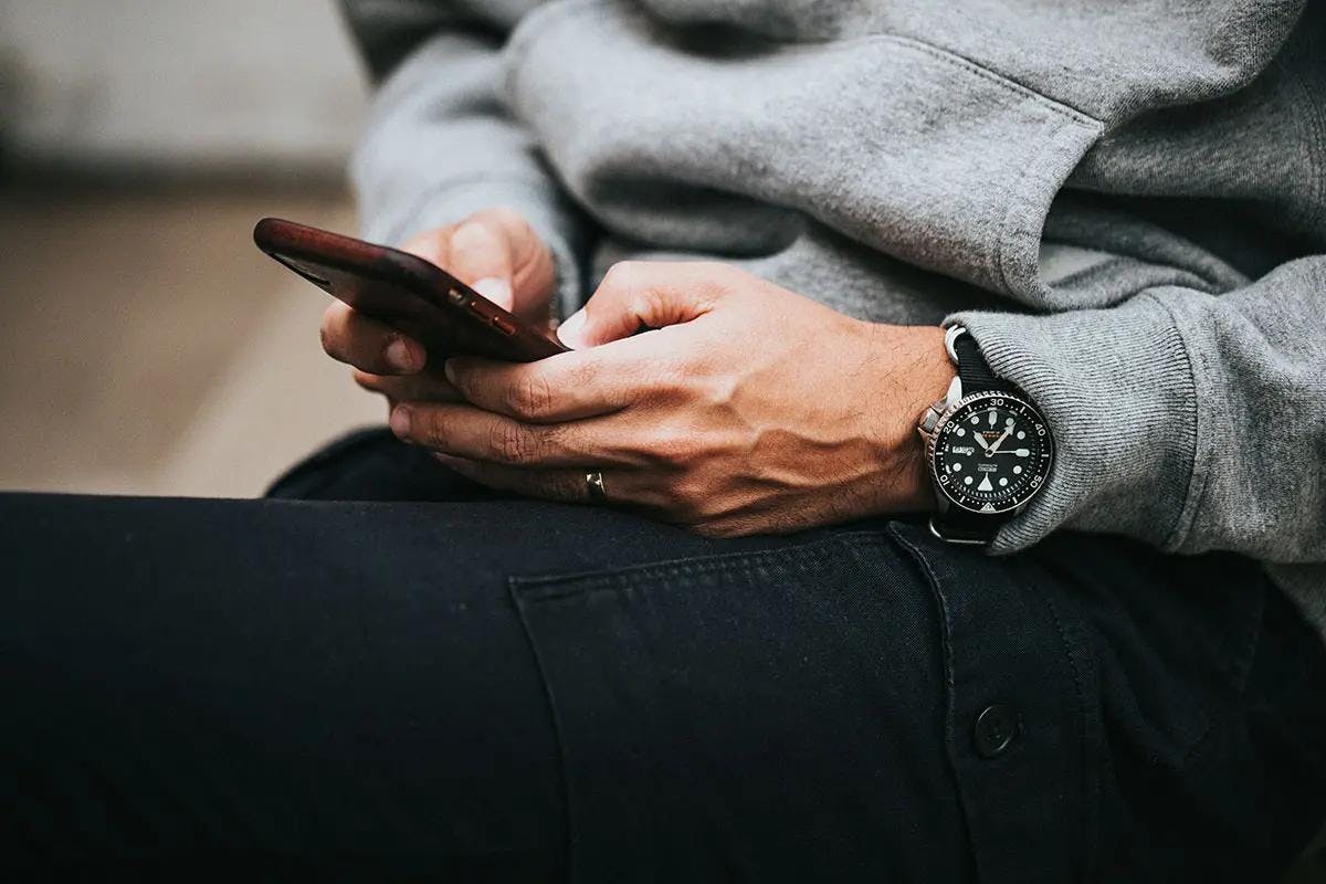 A close up image of a man on his phone wearing a grey hoodie, black jeans, and a black watch researching capital markets authority jobs.