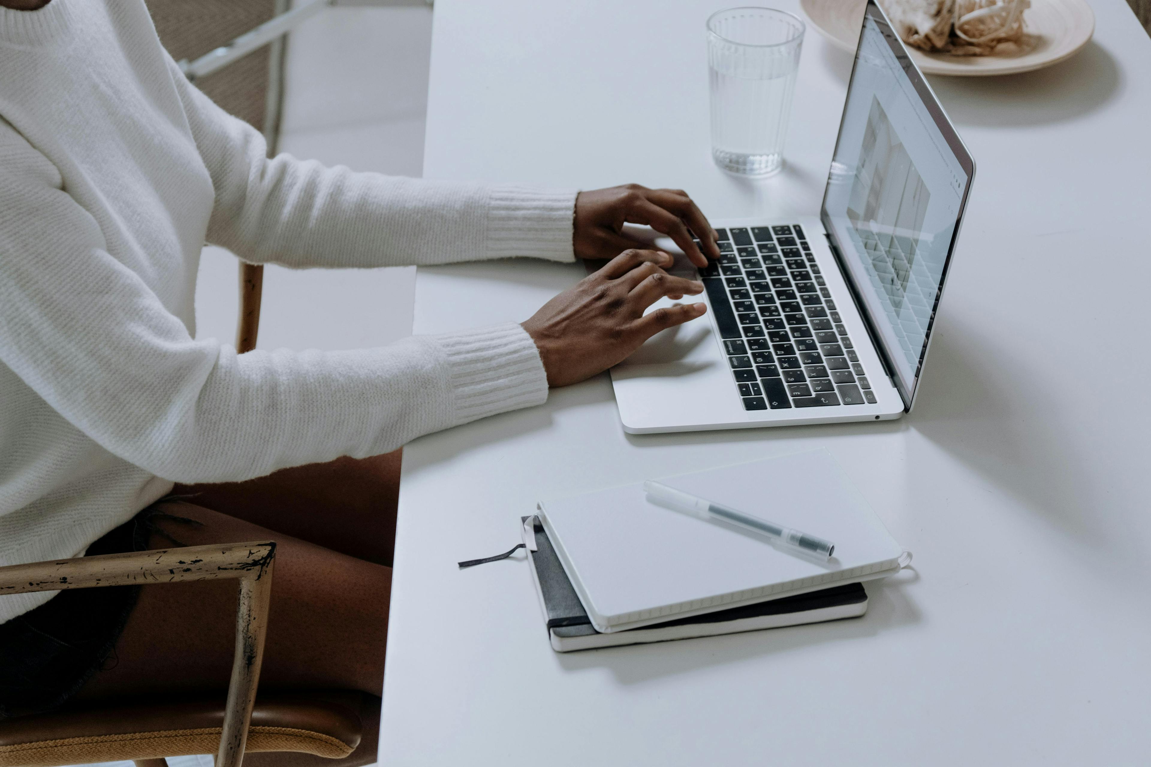 A person wearing a white sweater is typing on a laptop at a white desk. On the desk are a notebook, a pen, and a glass of water. The setting is minimalistic and clean. This image could represent a situation where the landing page experience is below average.