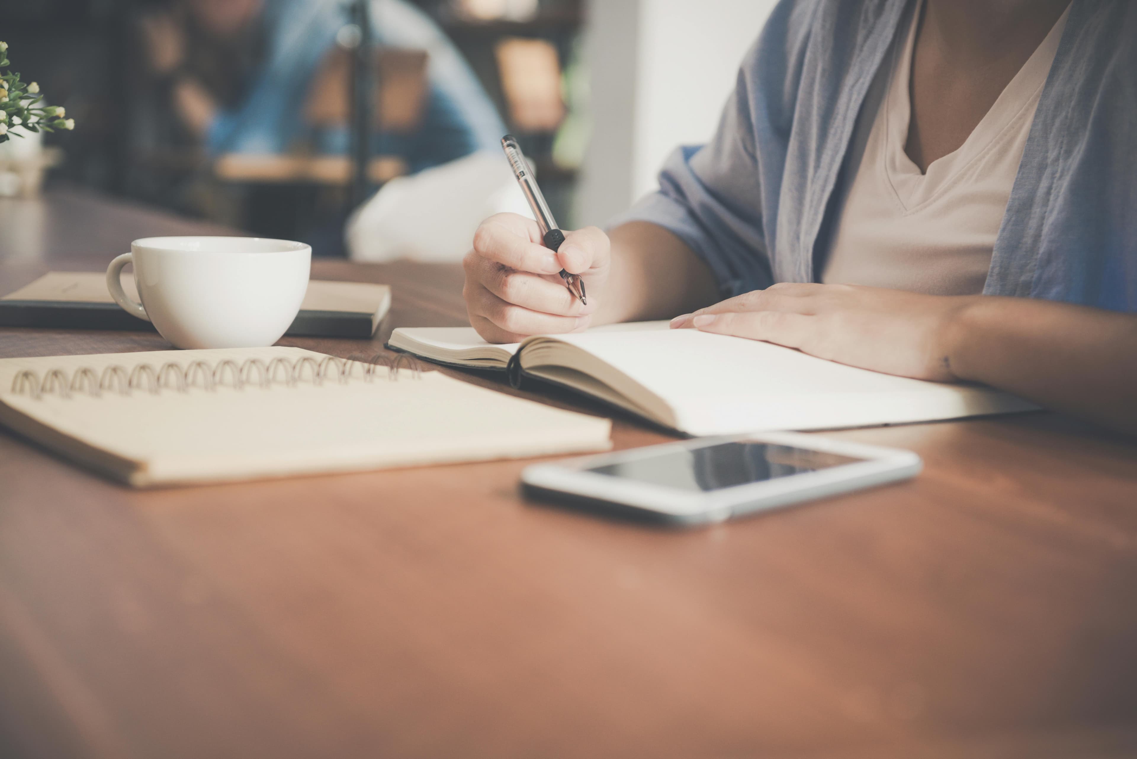 A person sits at a wooden desk, writing in a notebook with a pen, a coffee cup and smartphone nearby. Ideal for illustrating creative brainstorming or planning using an AB testing tool for content optimization.