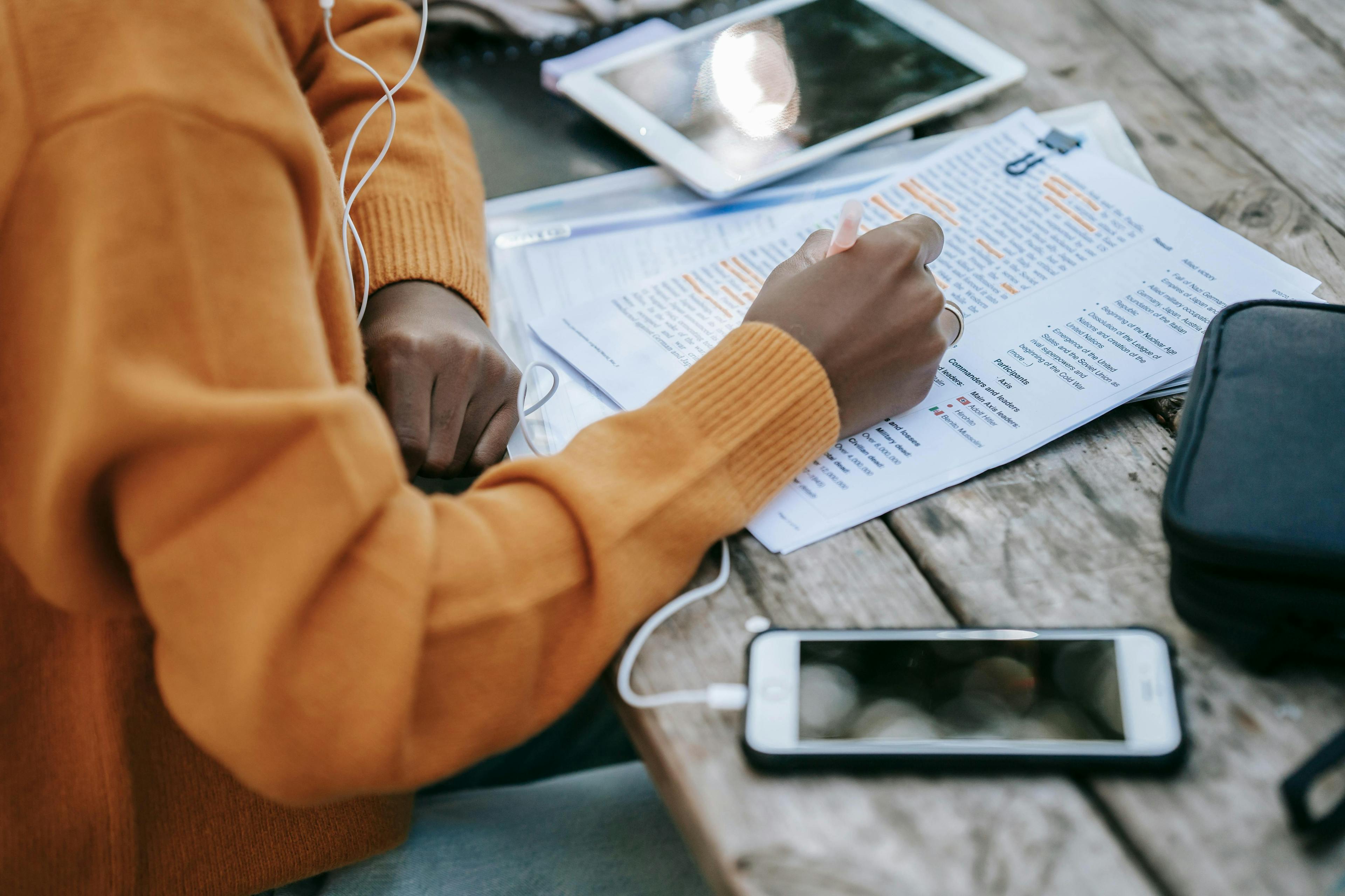 a person wearing an orange sweater, highlighting a text document with a marker, and listening to earphones. On the table are a tablet and a smartphone. This setting is perfect for exploring "lead generation services" to enhance business research and productivity.