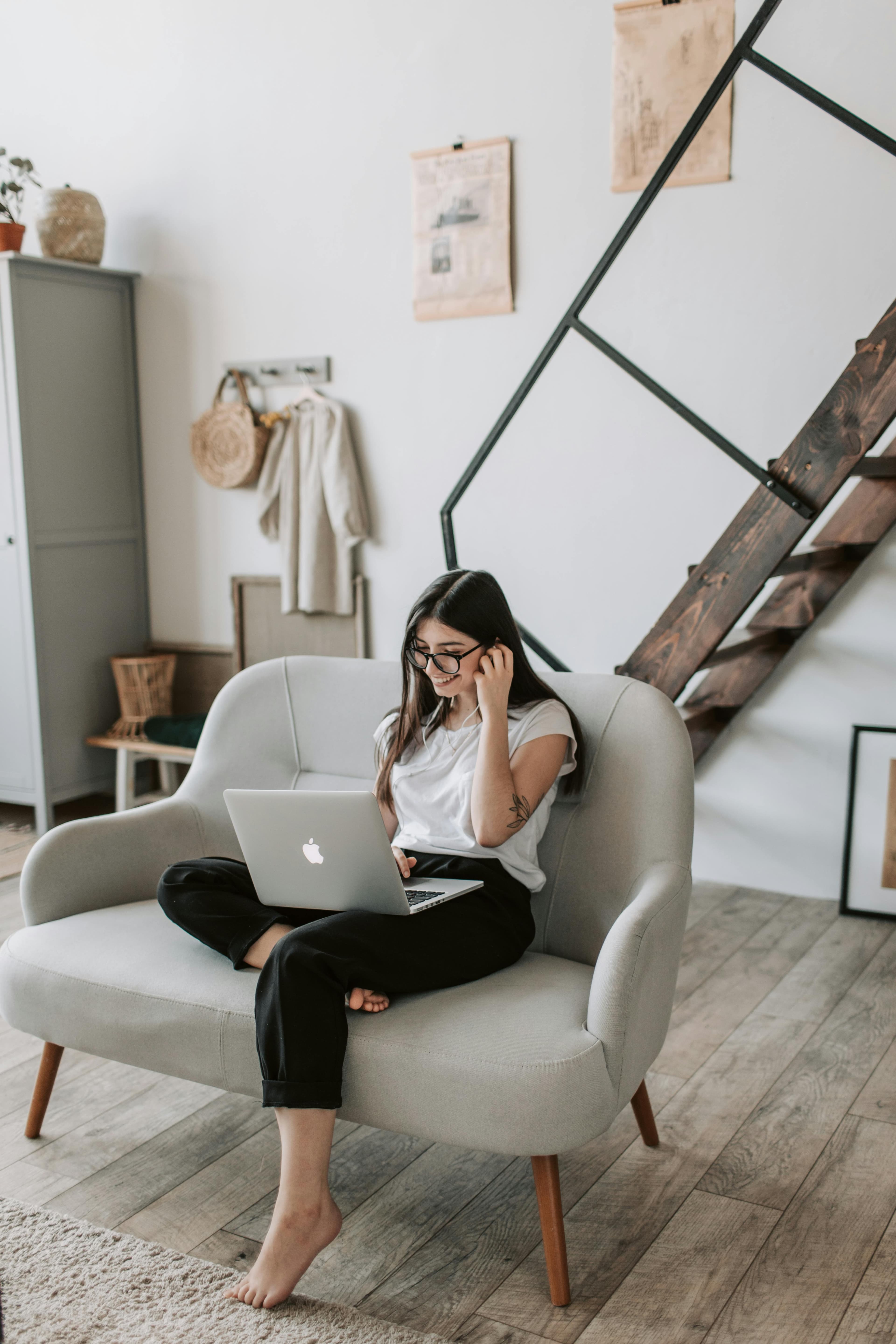 A woman sitting comfortably on a stylish sofa, using a laptop and wearing earphones. The light-filled room and serene atmosphere indicate that she could be conducting A/B testing software experiments, checking user engagement metrics or web optimization results.