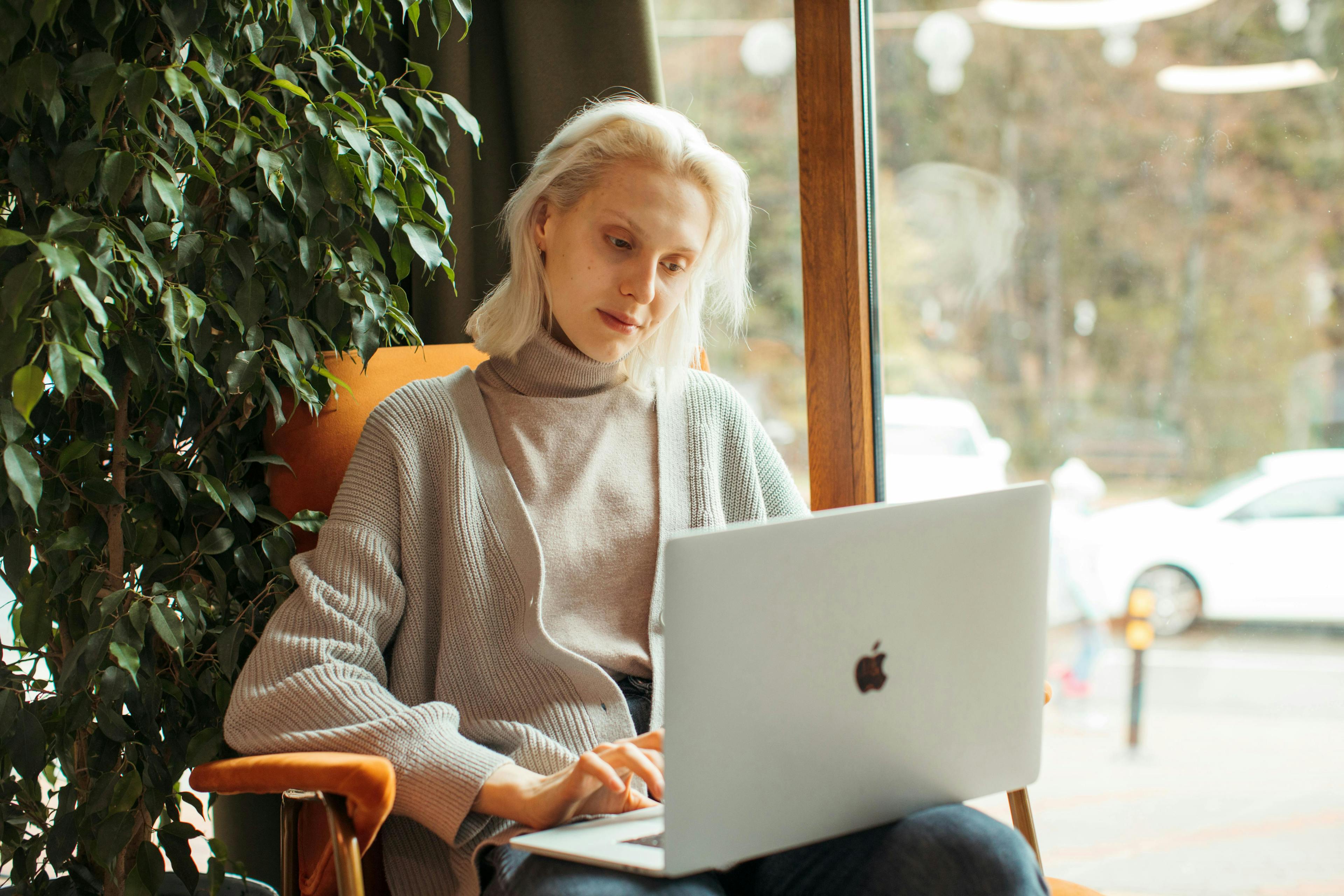A woman with blonde hair works on her laptop in a cozy chair by a window, surrounded by greenery. This image represents "homes for sale in Page, AZ."