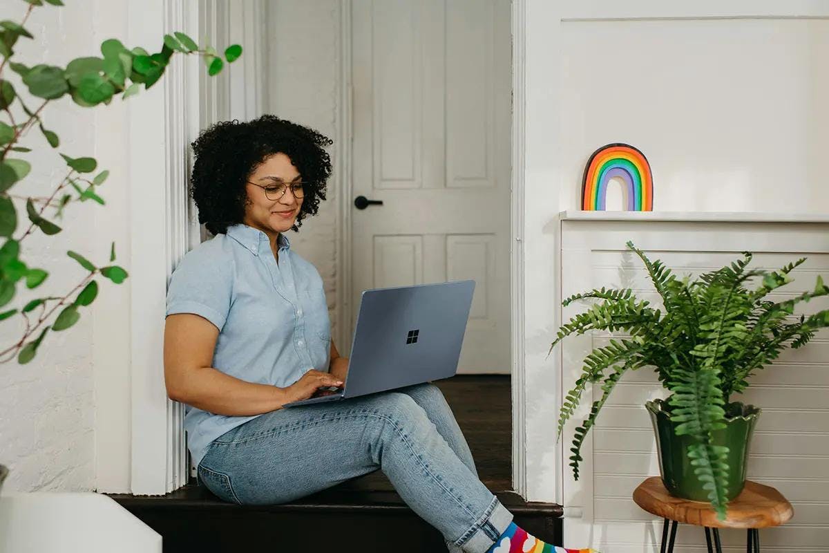 A woman with curly hair and glasses is comfortably seated on a bench, working on a laptop. She is wearing a light blue shirt and jeans, with colorful rainbow socks adding a playful touch to her outfit. The room is cozy, adorned with plants and a vibrant rainbow decoration on the shelf. The environment exudes a relaxed and creative vibe, perfect for "audio video marketing."