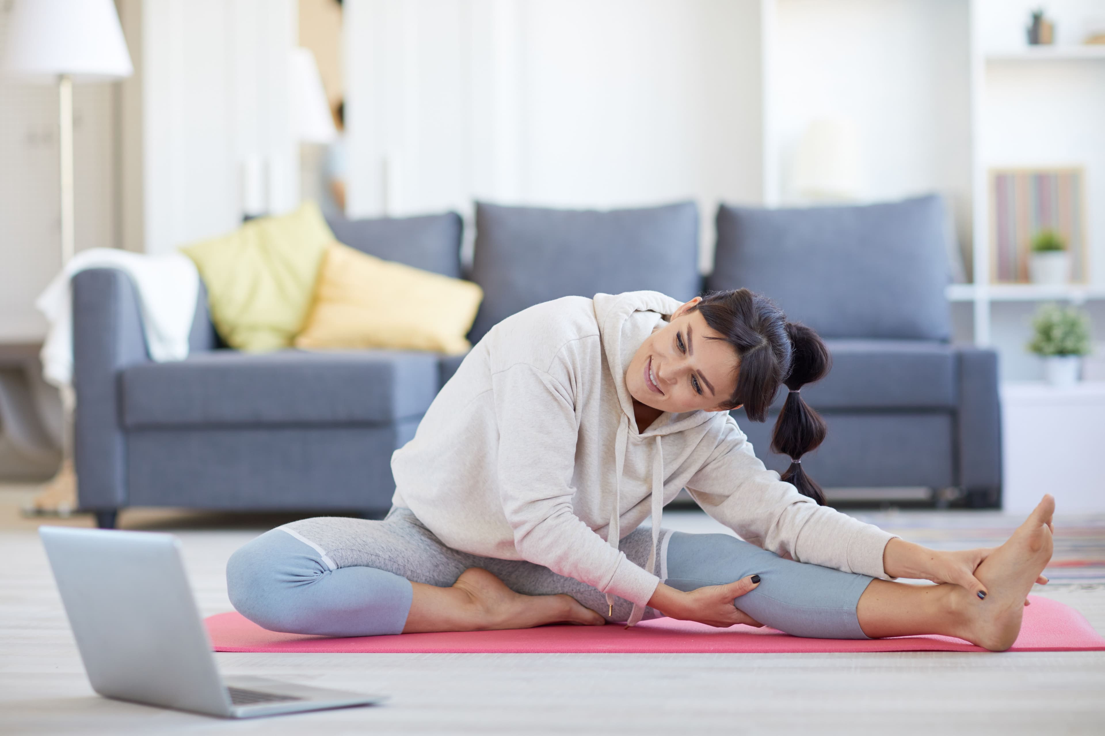 A woman stretching on a yoga mat in a bright living room, looking at a laptop screen. Ideal for small business websites offering online fitness classes or health and wellness coaching.