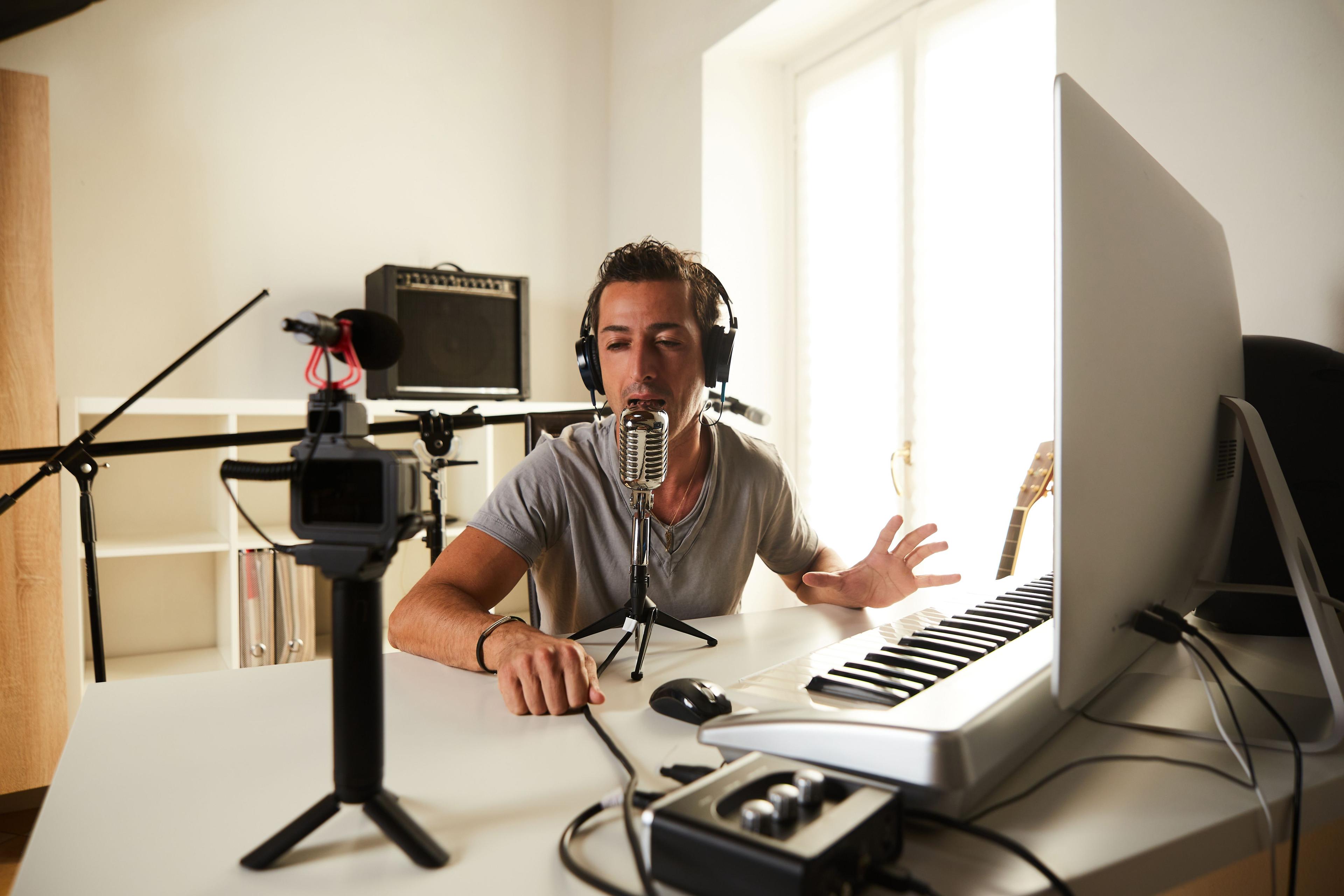 A man in a home studio speaking into a professional microphone, with headphones on, in front of a keyboard and computer. Recording equipment and a camera are set up, suggesting content creation or podcasting. Keywords: train_split_test, audio recording, content creation.