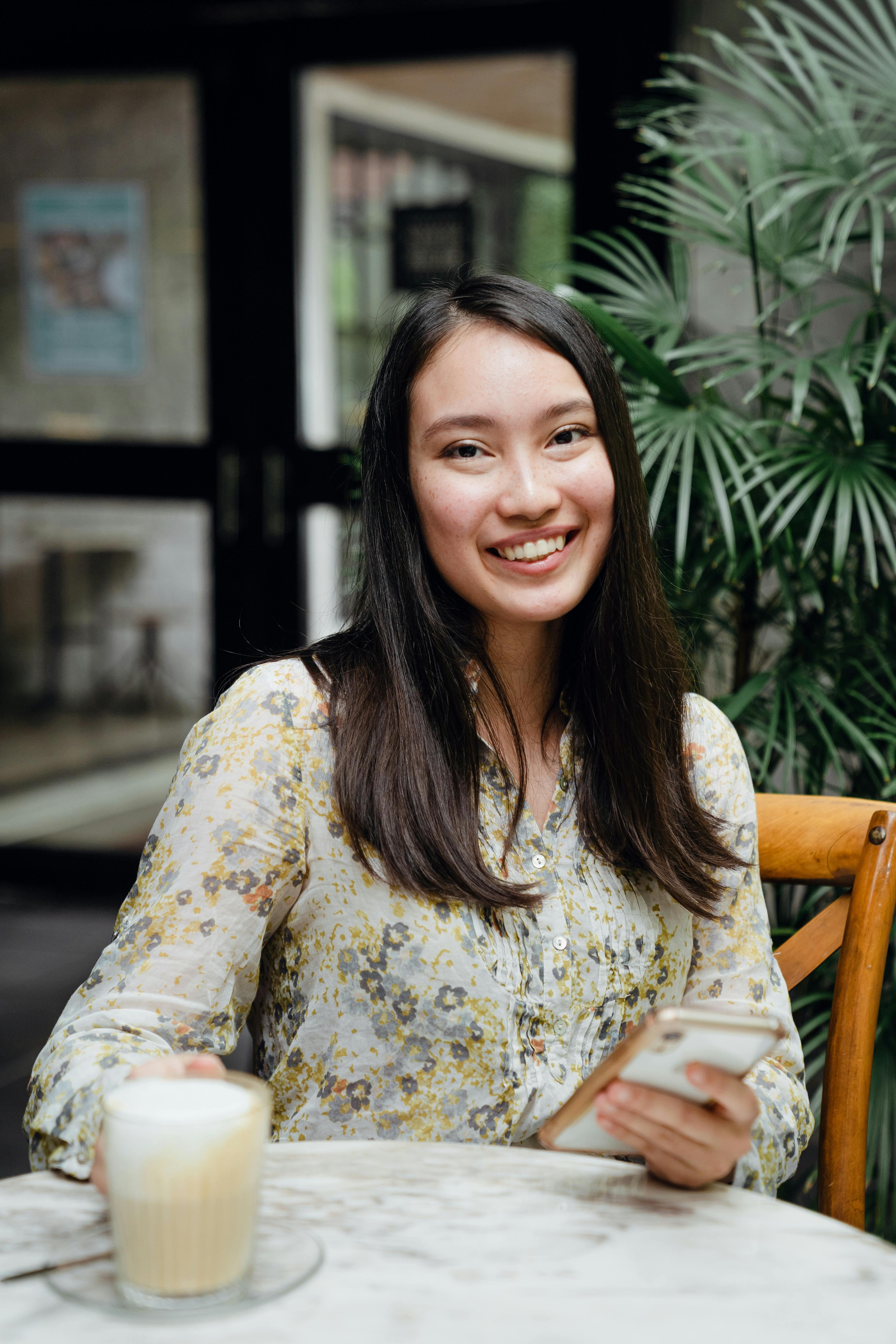 A woman smiling while holding a smartphone and enjoying a coffee at a café.