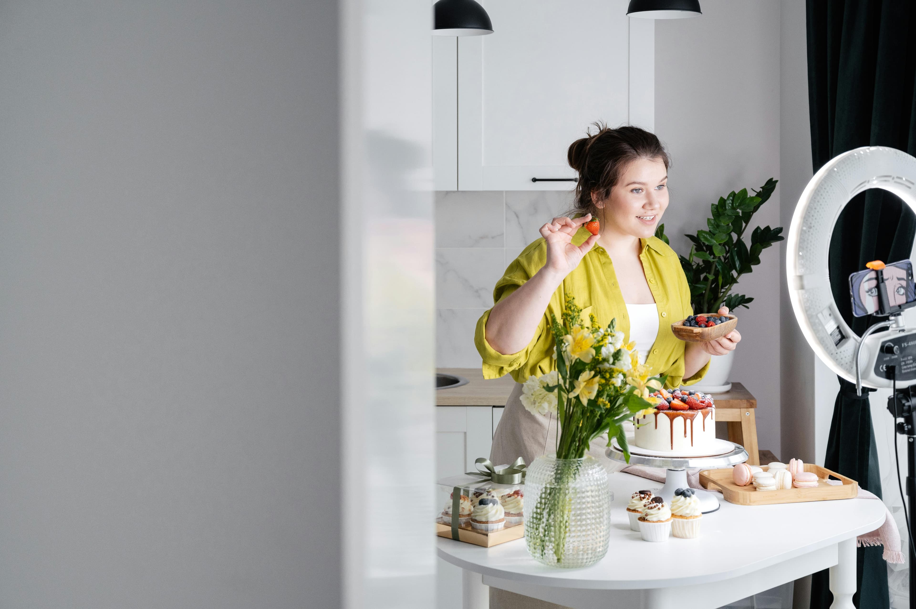 "A woman in a bright kitchen setting, presenting a beautifully decorated cake and berries to the camera, with a ring light and smartphone setup for recording, illustrating how engaging visuals on websites that convert can enhance audience interaction and sales.