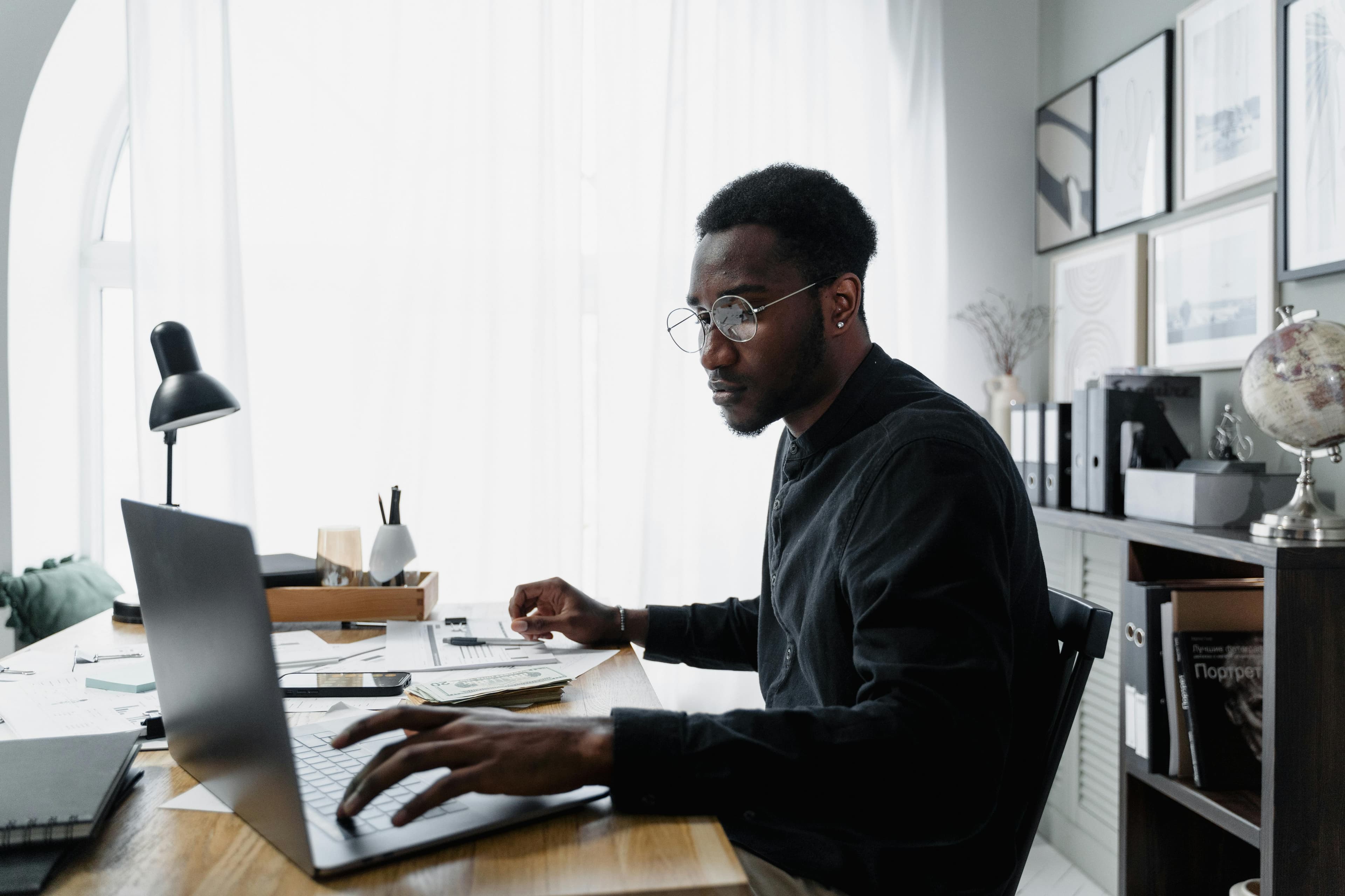 A man with glasses works intently on his laptop at a well-lit home office, surrounded by documents. This image highlights focus and productivity, perfect for showing someone using an AB testing tool to analyze data or run experiments.