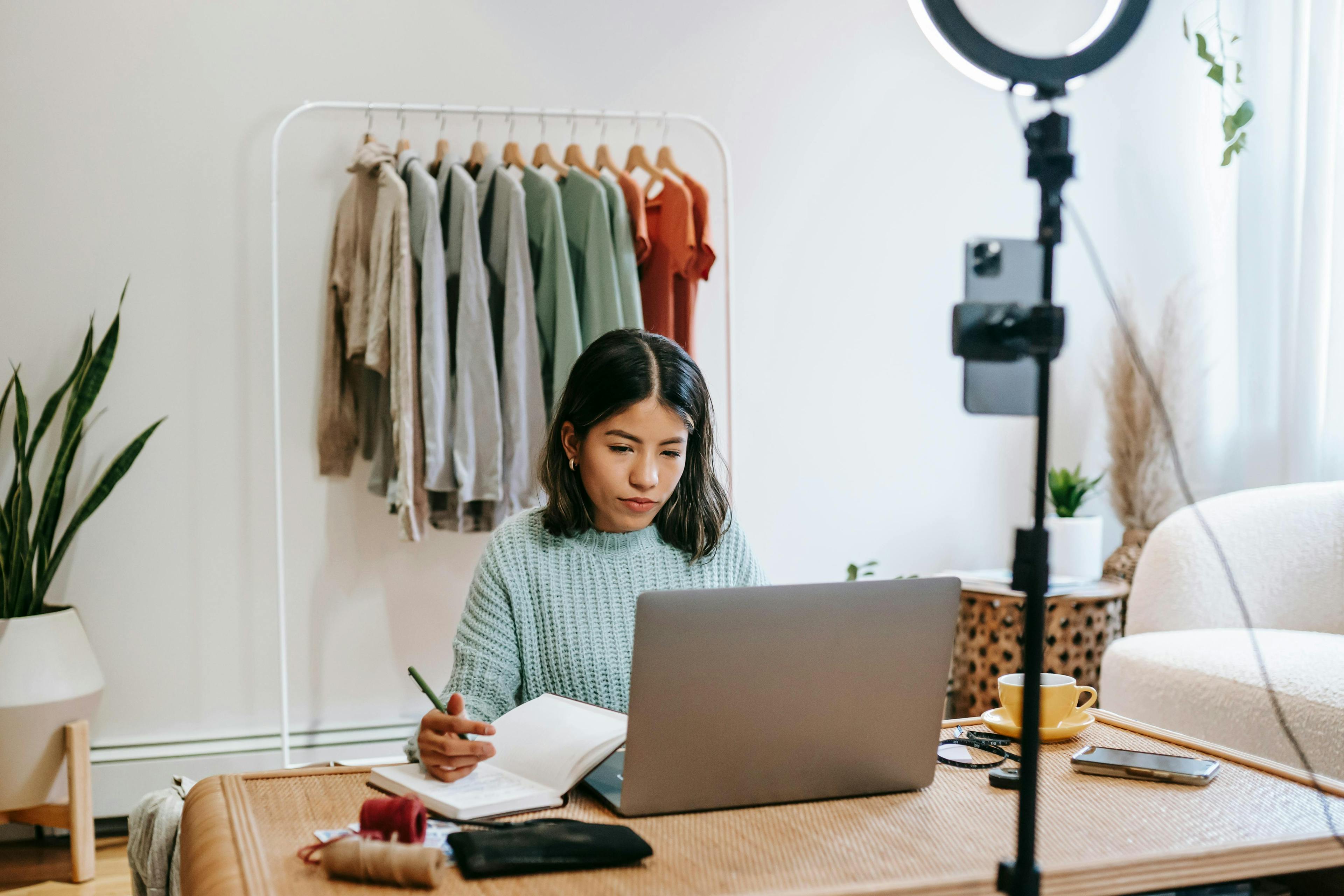 A young woman in a blue sweater taking notes while working on her laptop in a stylish home office setup, preparing for market events this week.