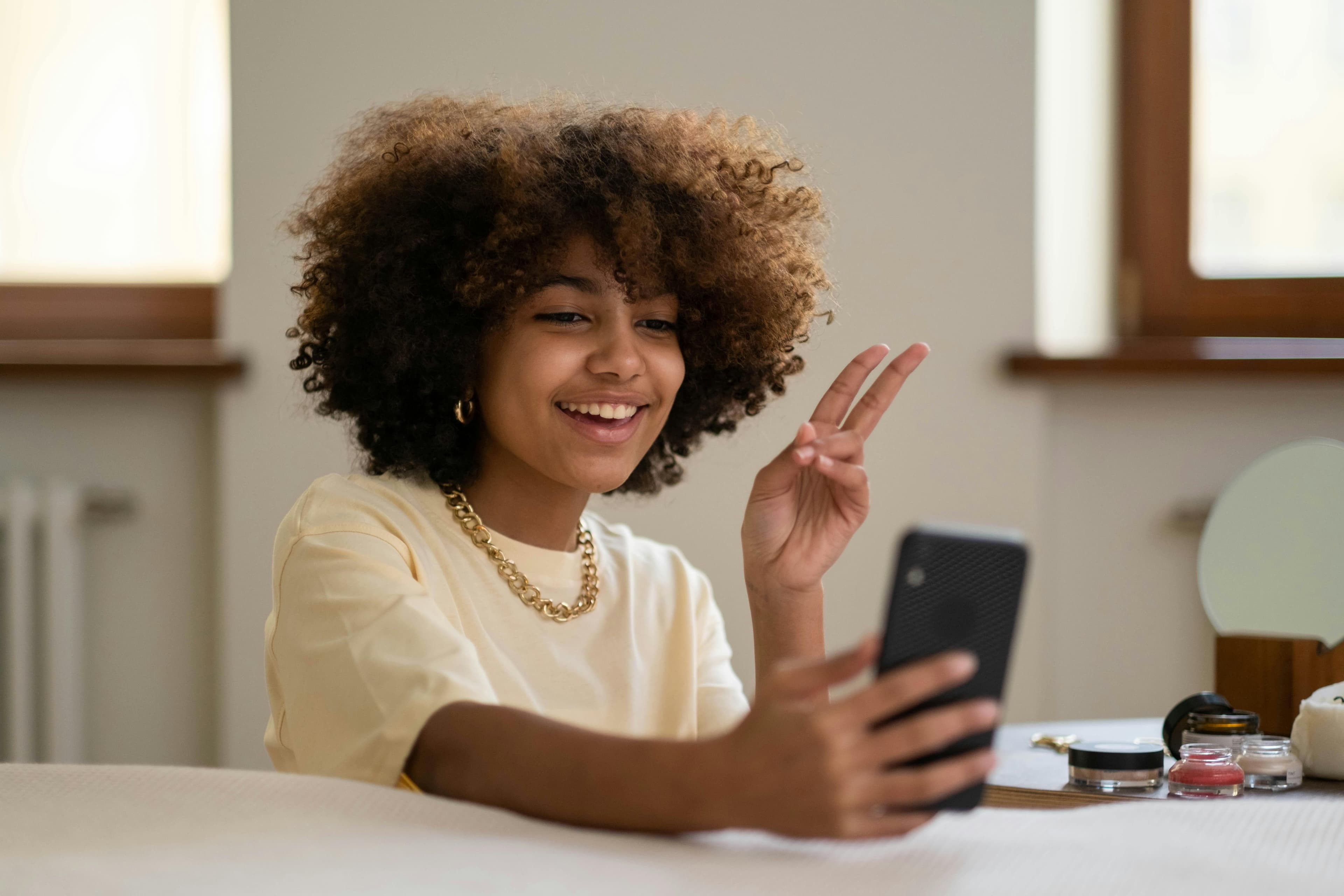 A young woman with curly hair smiles and gives a peace sign while recording herself on a smartphone. This scene illustrates the concept of "what is content marketing" by showcasing how individuals create engaging, personal content to connect with audiences through social media.