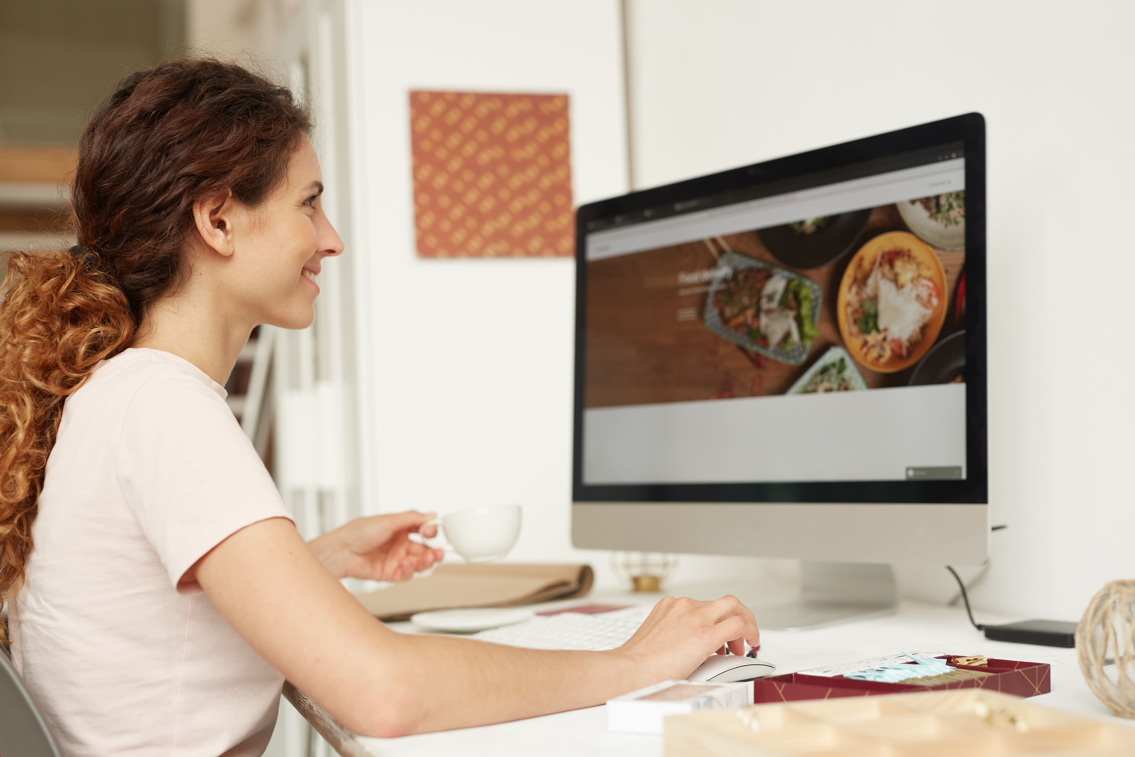 A woman sitting at a desk, smiling while holding a coffee cup and working on a desktop computer. The screen shows a food-related website, suggesting web design or content development. This image reflects the personalized guidance of a "small business marketing coach" helping clients build or enhance their online presence.