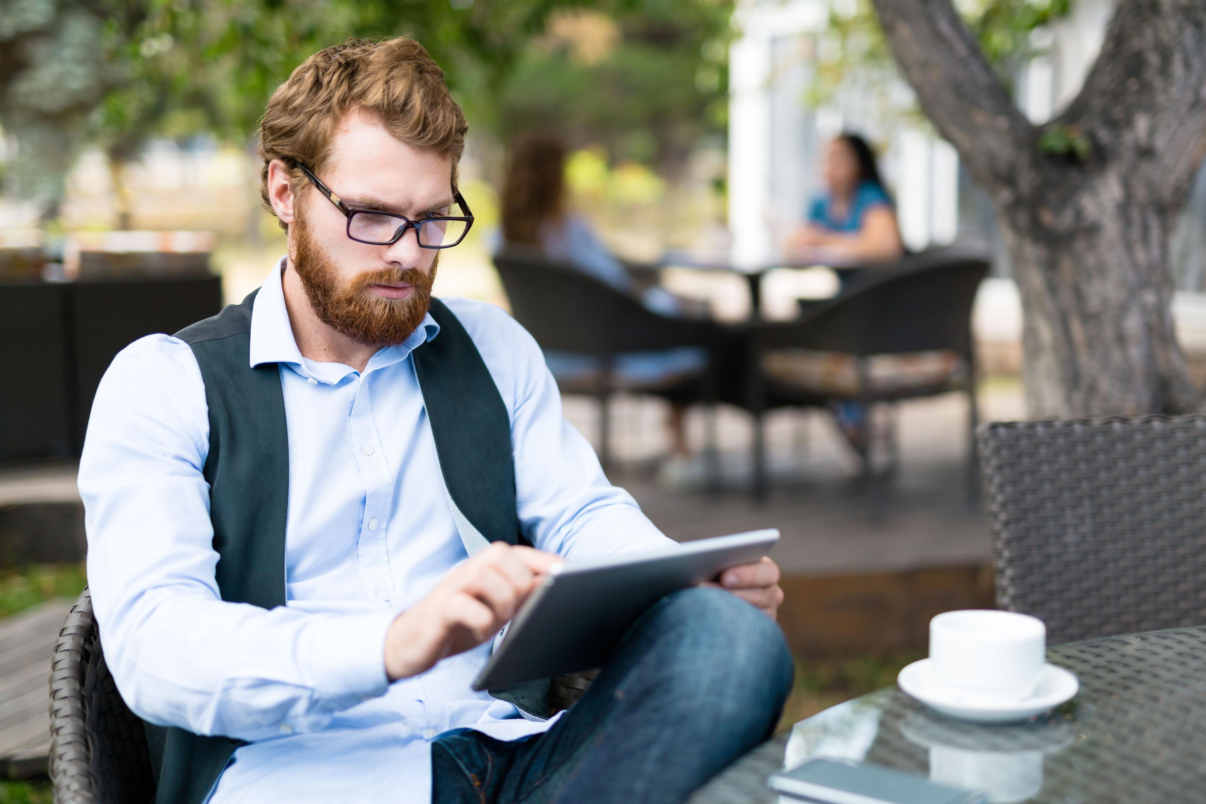 A man with a beard and glasses is seated outdoors, using a tablet to analyze data and track conversion calculator metrics while sipping coffee in a casual business setting.