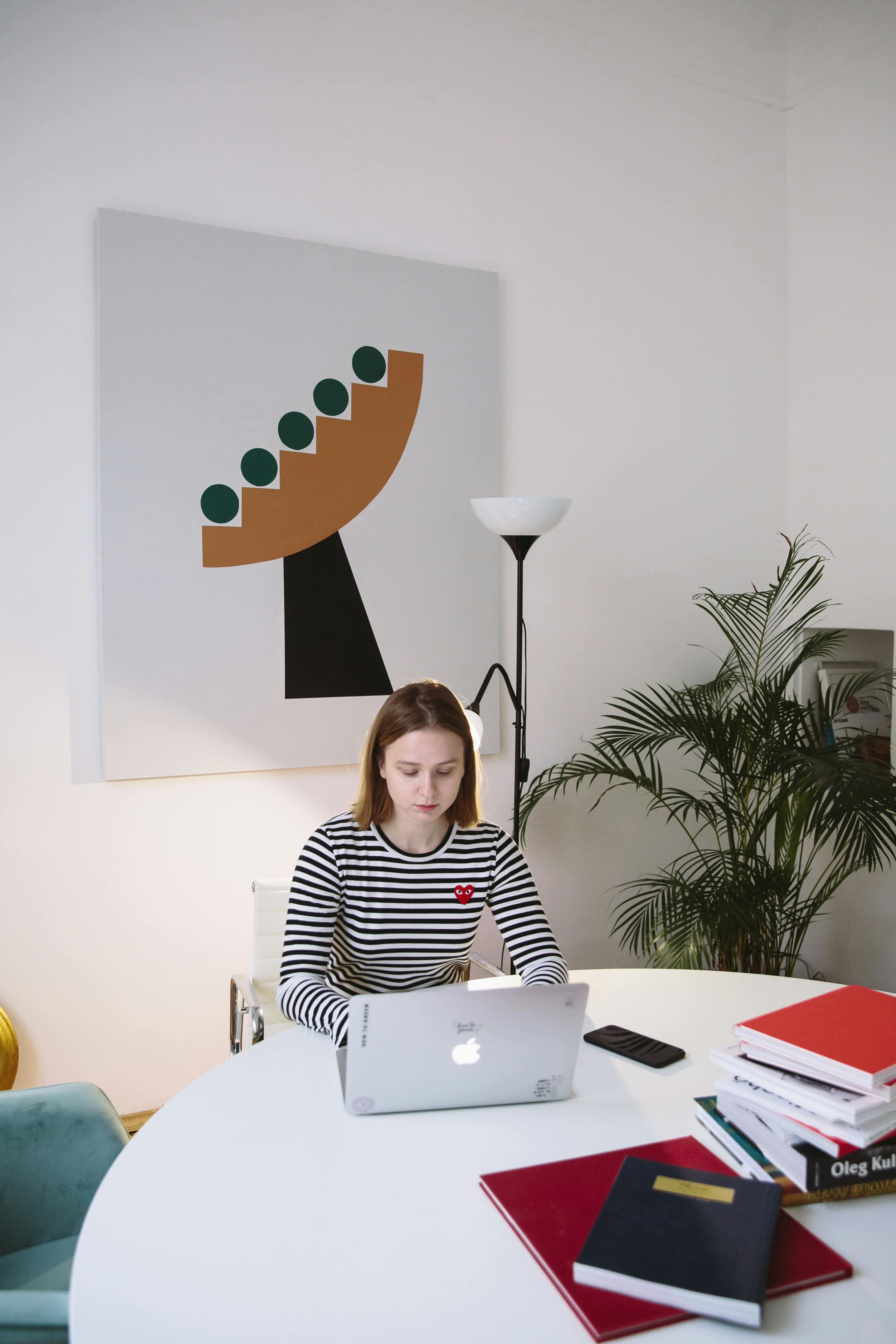 A young woman sits at a modern, minimalist workspace, working on her laptop with focus. The white walls and geometric artwork in the background create a clean, professional setting. This image could represent someone analyzing results from a split testing experiment, a method used to compare two versions of a webpage or product to determine which performs better.