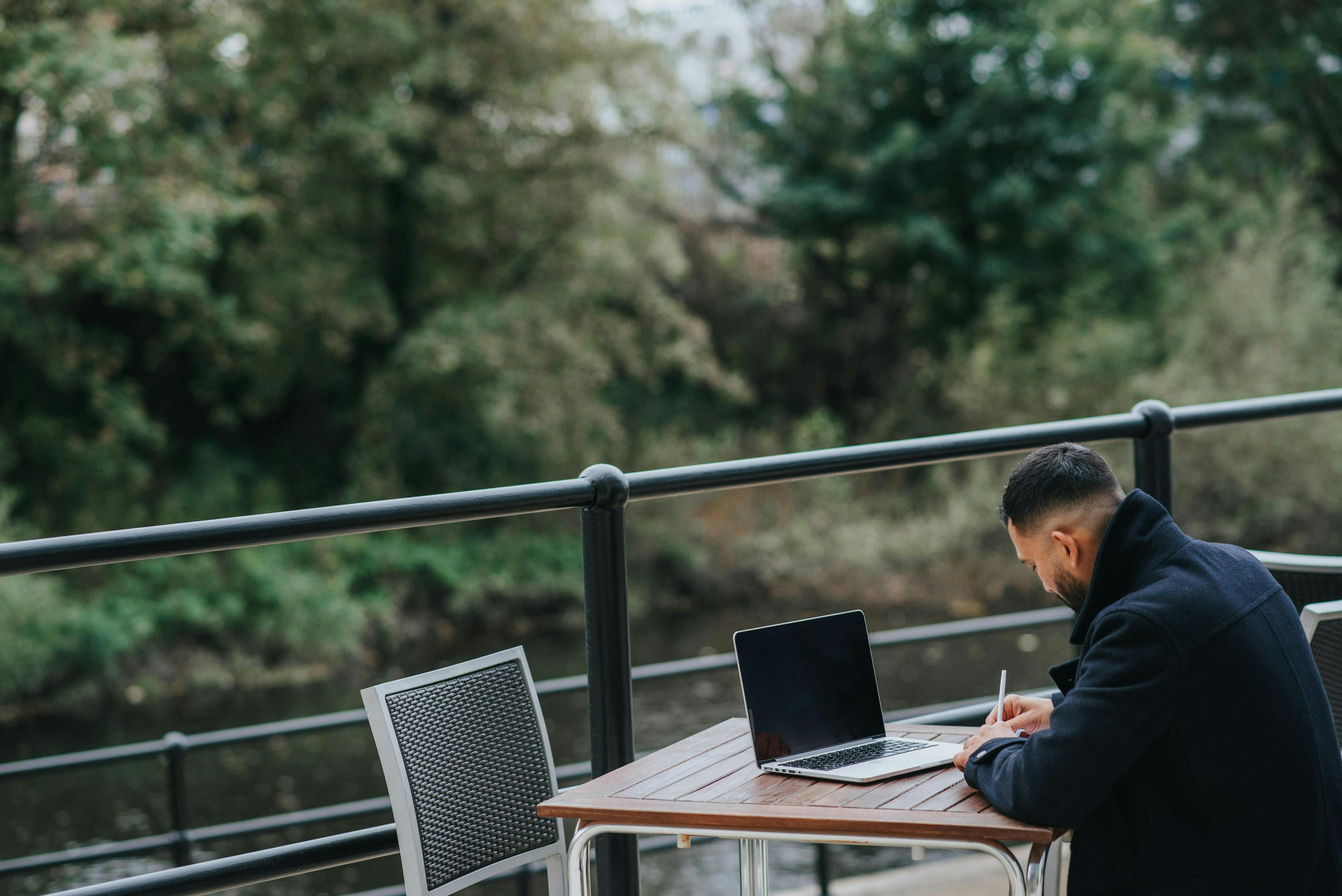 A man in a dark coat sits at an outdoor table near a river, working on his laptop and writing in a notebook. The setting is peaceful, with trees in the background. This image represents "high point marketing authority."