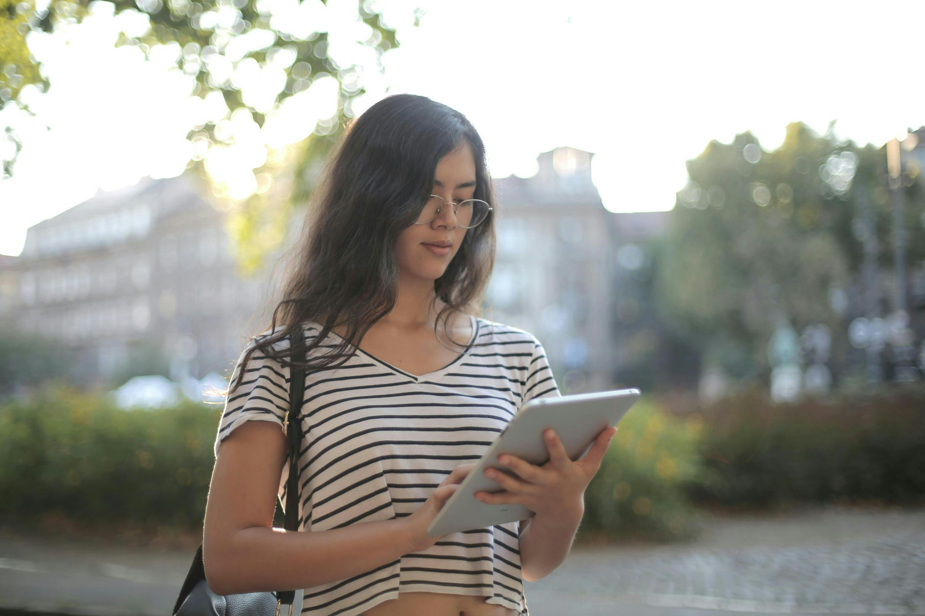 A young woman in glasses and a striped shirt stands outdoors, using a tablet, representing a digital marketing company.