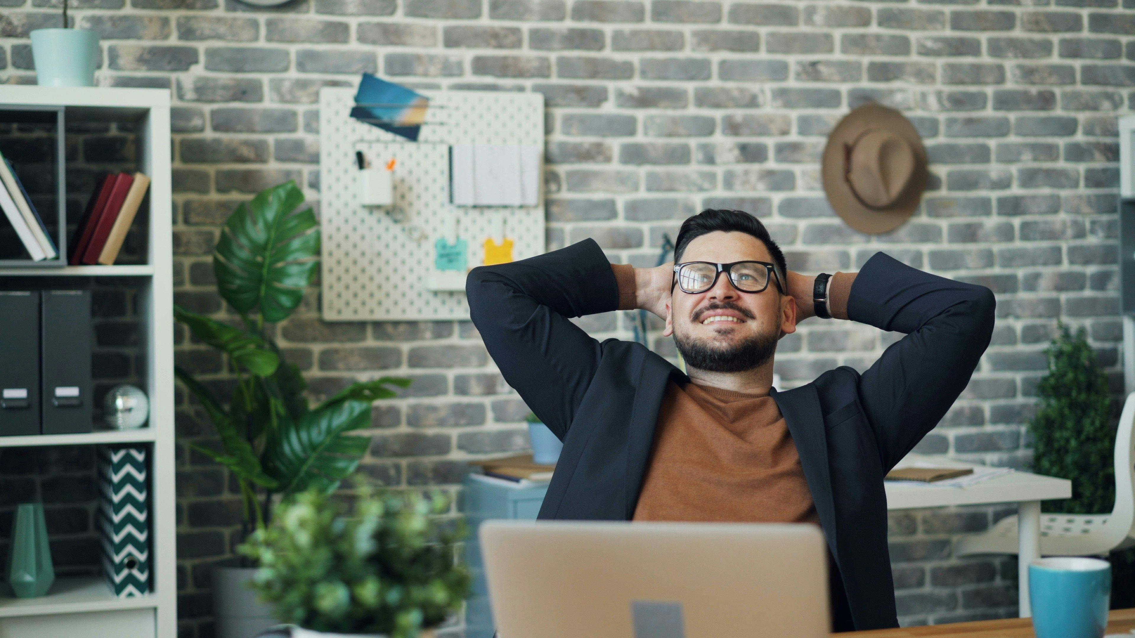A man in a stylish office leans back in his chair with a satisfied smile, looking relaxed and confident. The modern workspace features a brick wall, indoor plants, and a laptop on the desk, suggesting productivity and creativity. This image is ideal for illustrating the ease and benefits of AI copywriting tools available for free.