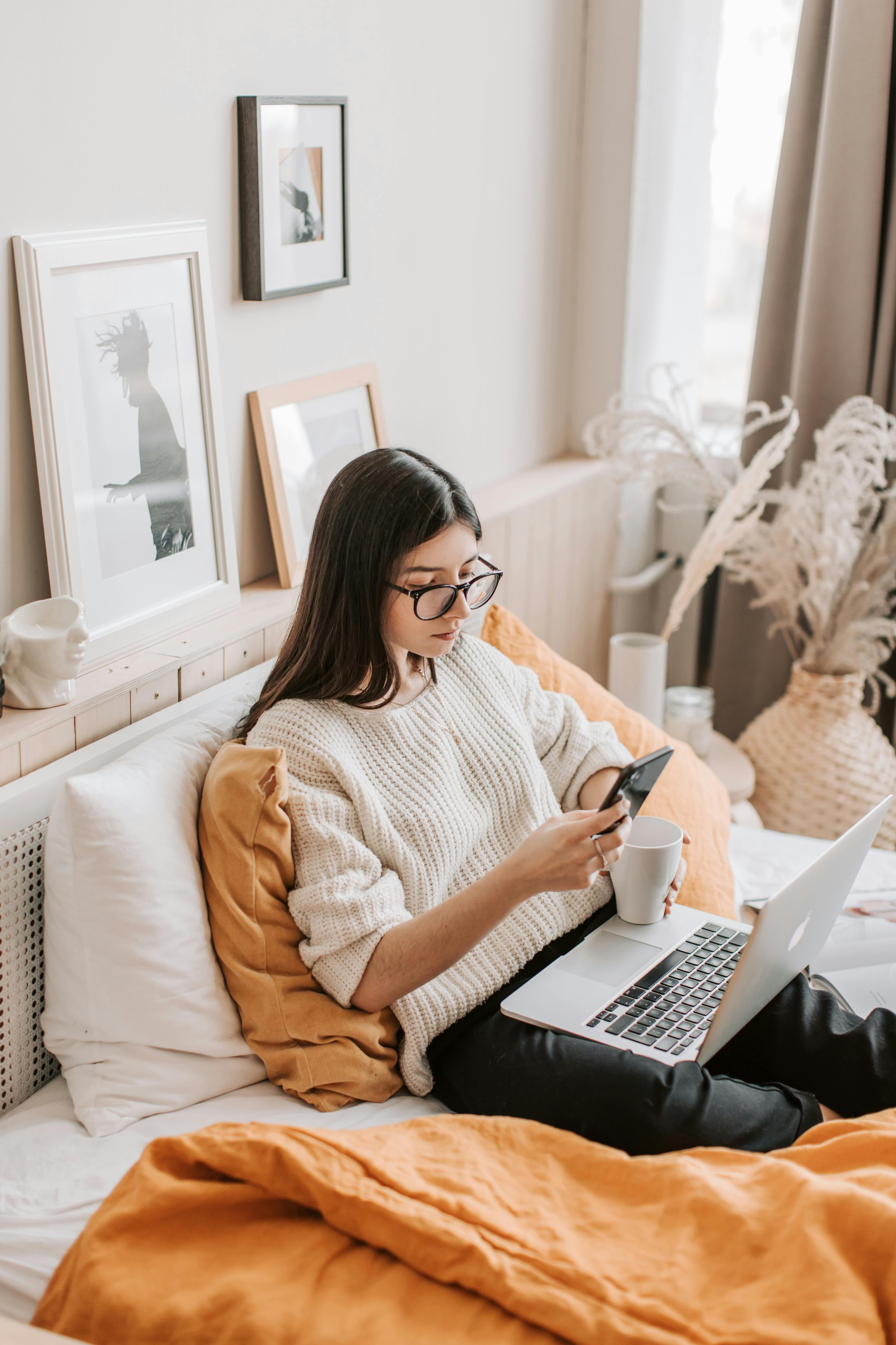 A woman sitting on her bed with a laptop and smartphone, using an A/B testing program to evaluate marketing strategies.