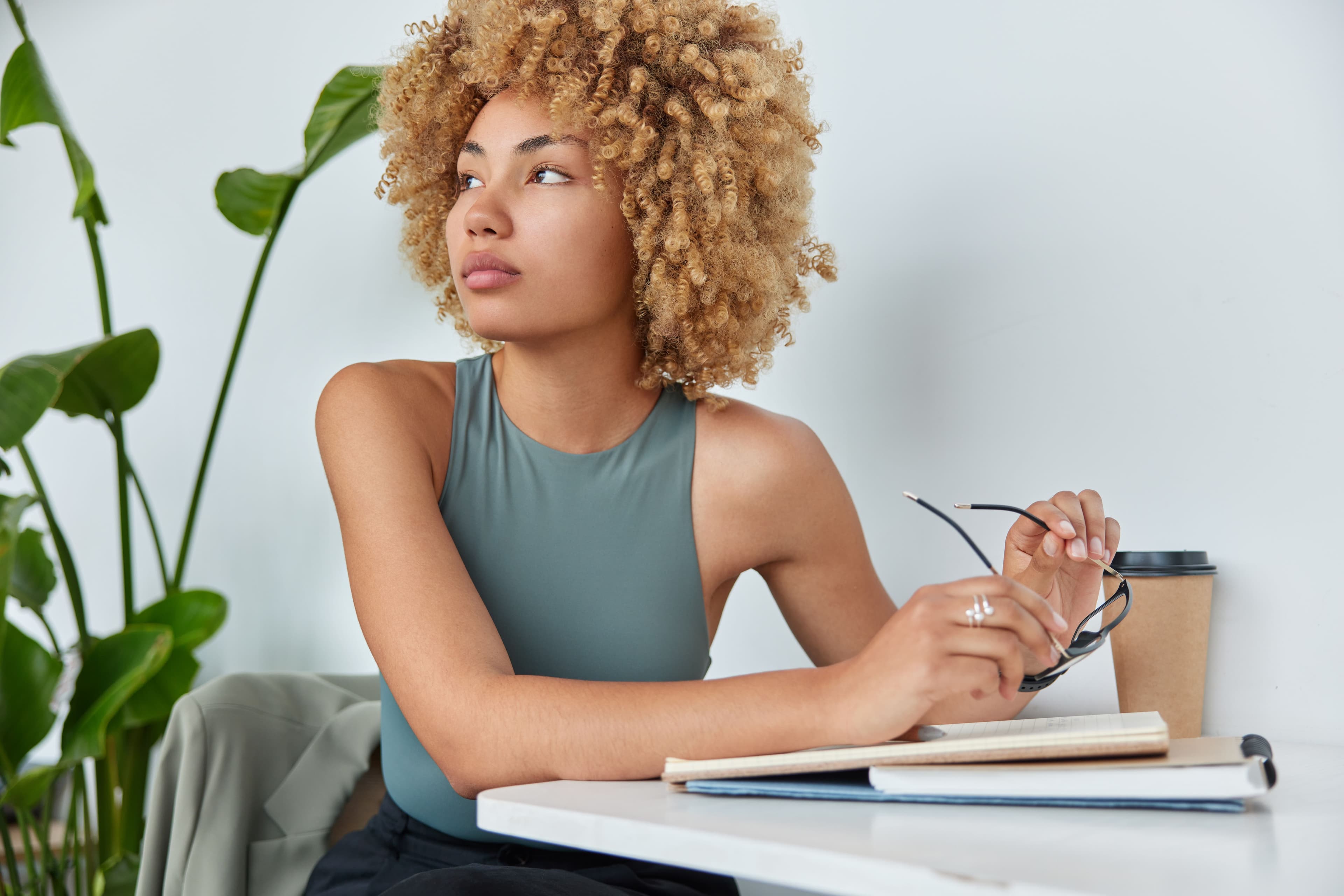 A young woman in a thoughtful pose, holding her glasses with a notebook and coffee nearby, capturing the essence of creative brainstorming in a "social media ad agency."