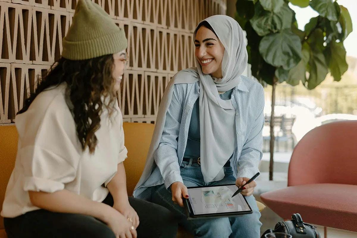 Two women are sitting on a yellow bench, engaged in a friendly and animated conversation. One woman, wearing a hijab and a striped shirt, is holding a tablet and pointing at the screen with a stylus. The other woman, in a beanie and a light sweater, is listening attentively and smiling. The background features a stylish wall and greenery, creating a modern and inviting atmosphere. This scene is ideal for illustrating the concept of a "B2B SaaS marketing agency."