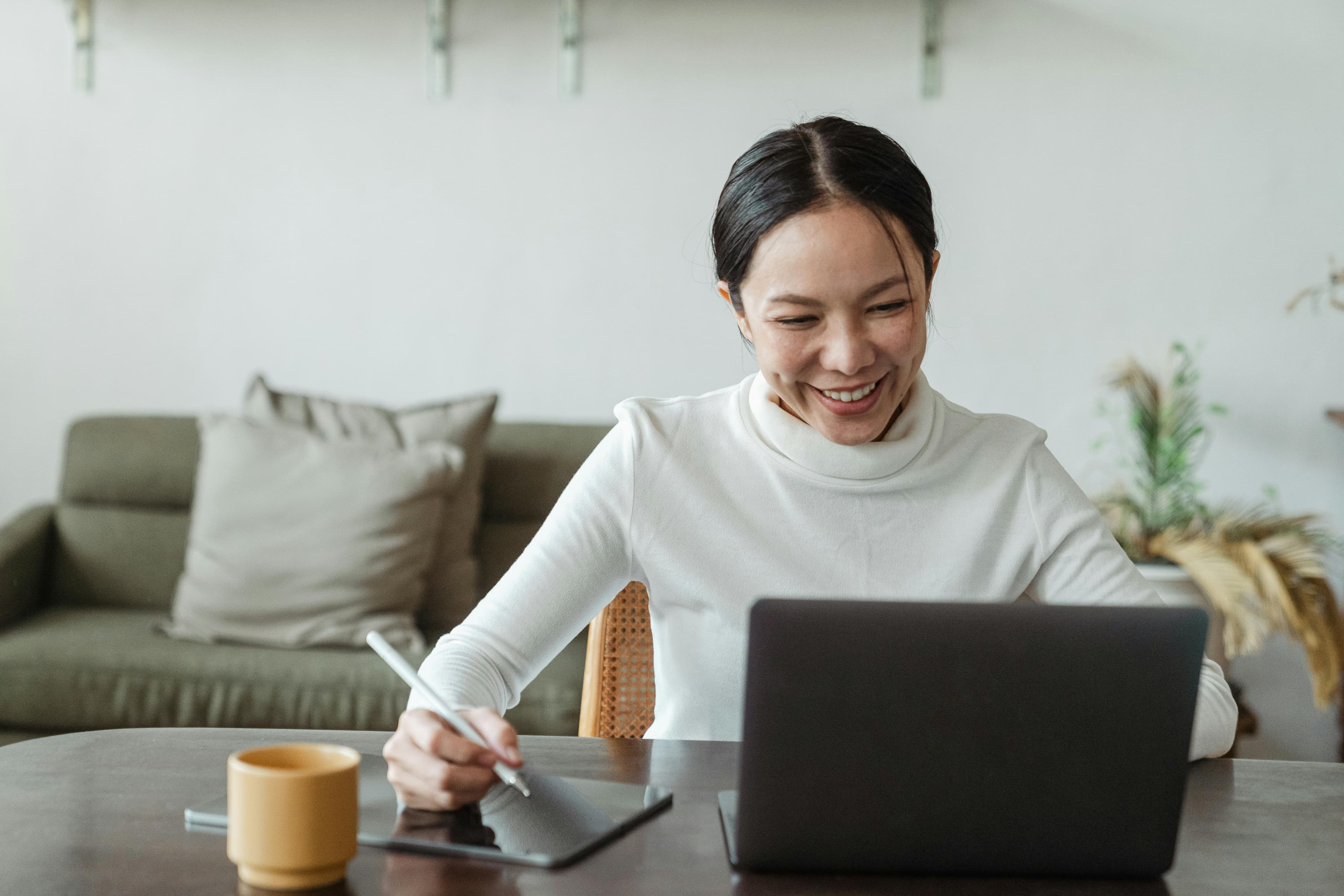 A smiling woman sits at a table, working on a laptop. She holds a stylus and is using a tablet beside her computer, indicating she might be involved in design or digital marketing tasks. On the table, there is also a yellow mug. The cozy background includes a sofa with cushions and decorative plants, creating a comfortable workspace. This image portrays someone engaged in full funnel marketing, planning and executing strategies to guide potential customers through the entire buying journey from awareness to conversion.