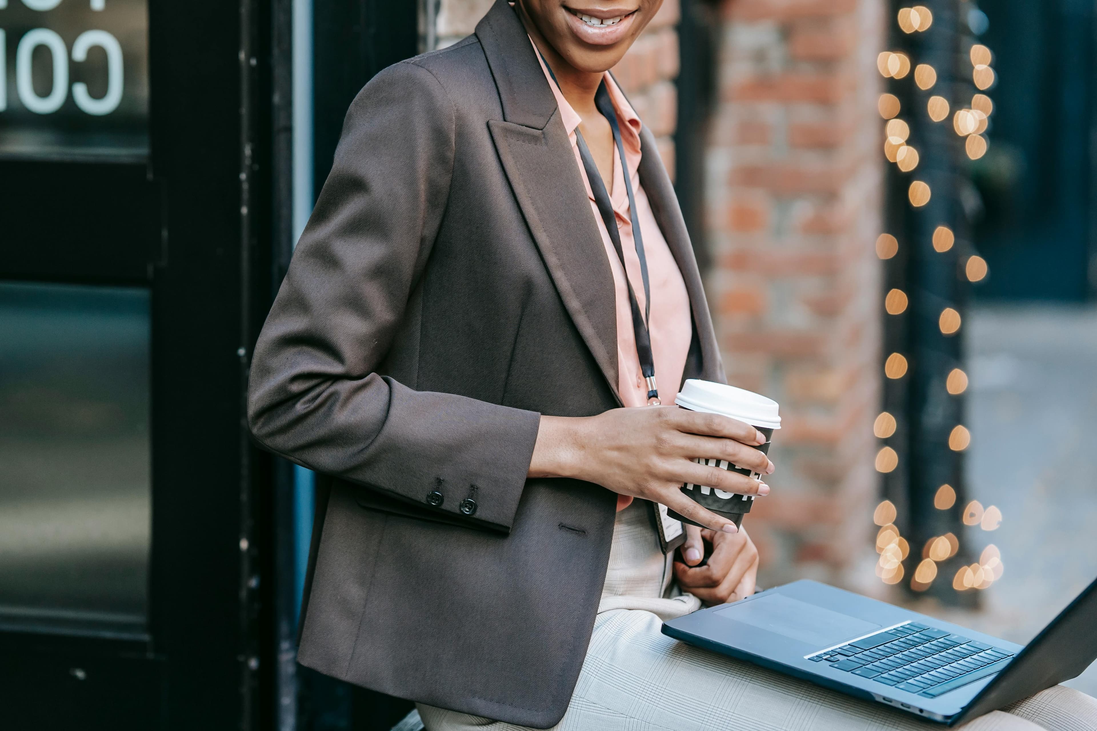 A woman in a professional outfit, wearing a brown blazer and holding a coffee cup, sits outdoors with a laptop on her lap. She is smiling, suggesting a positive and productive work session. The background features a brick wall adorned with twinkling lights, adding a festive touch. This image likely depicts someone working on an event marketing plan, taking a moment to enjoy a coffee break while strategizing and organizing event details.