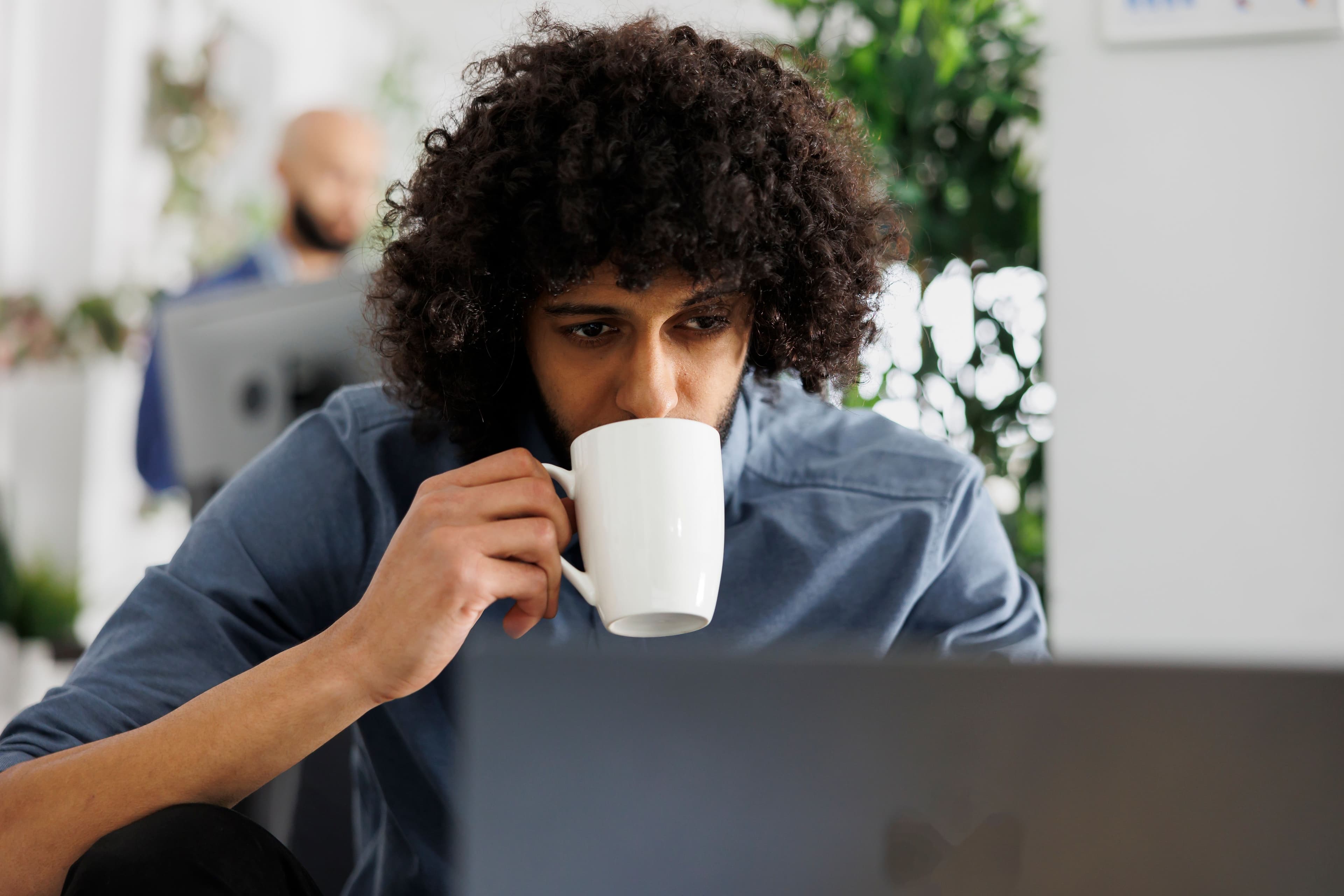 A man with curly hair, wearing a blue shirt, is intently looking at a laptop screen while sipping coffee, possibly checking the stock market performance today in a busy office environment.