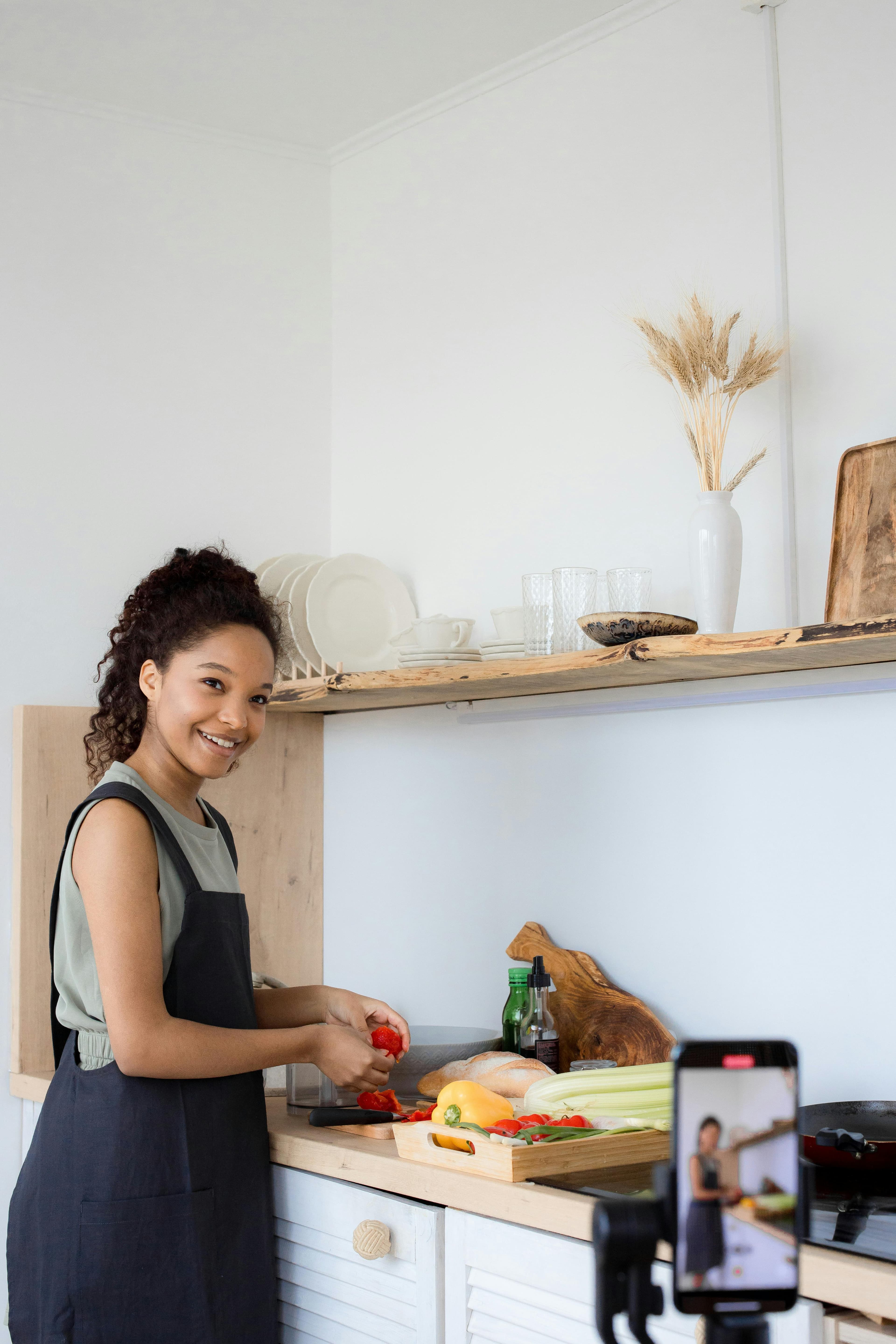 A smiling woman preparing fresh vegetables in a kitchen while recording a video with a smartphone. This setup answers the question "what is a lead magnet" by demonstrating how video content, such as cooking tips, can be used to attract and engage an audience online.