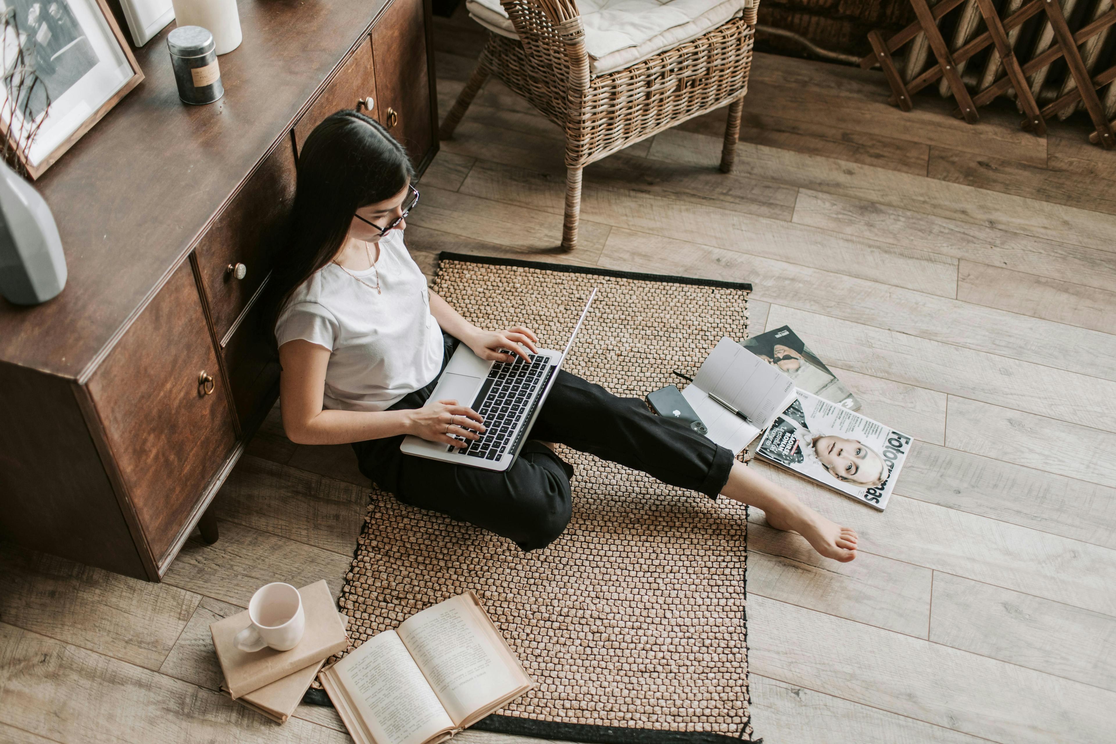 A woman working on her laptop while sitting on a rug in a cozy room, surrounded by books and magazines. This image is ideal for promoting lead management software that enhances productivity and organization in a home office setting.