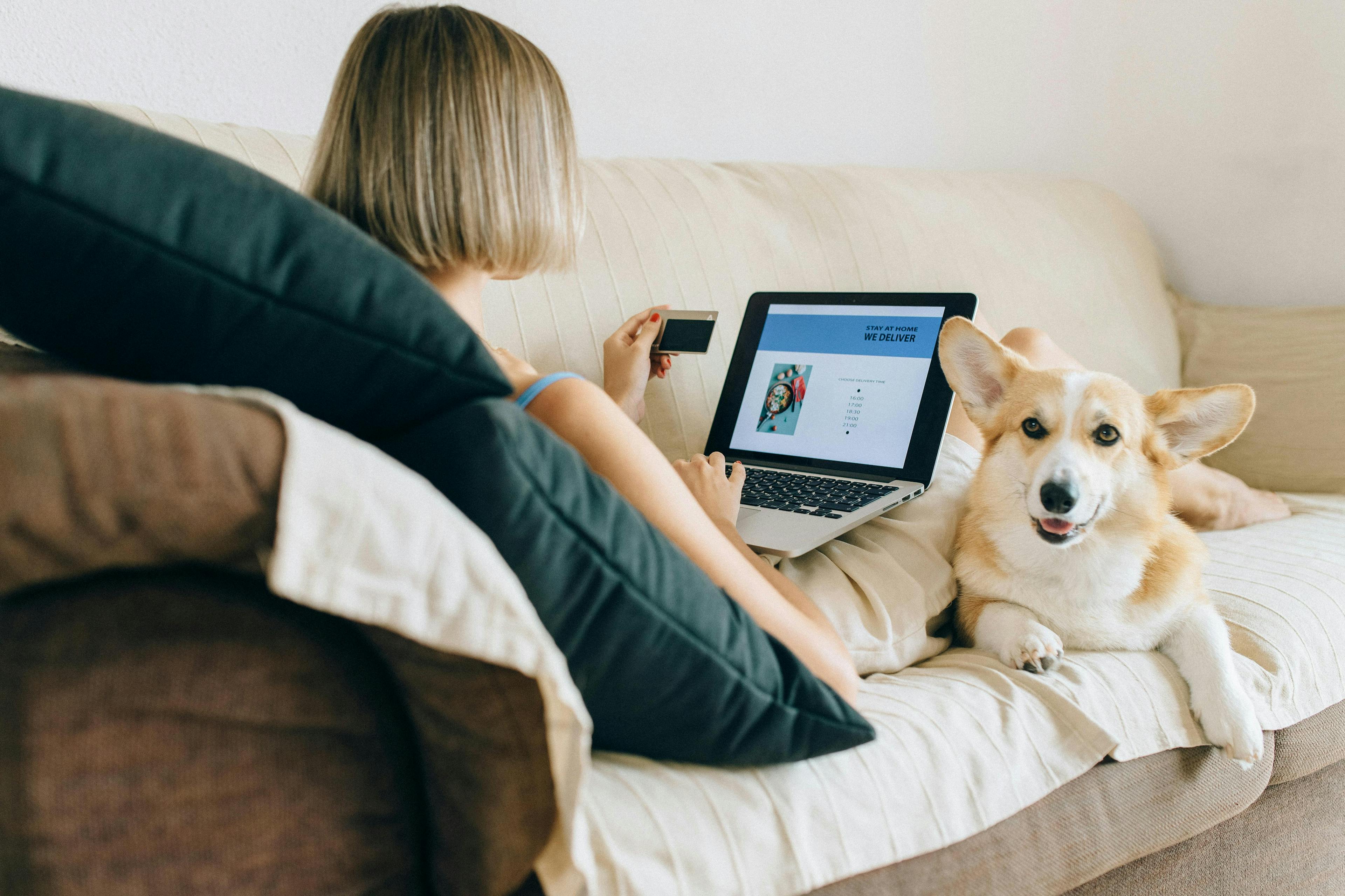 A woman lying on a couch with a laptop on her lap, holding a credit card. Next to her is a smiling corgi dog. The laptop screen displays an online delivery service with the text "STAY AT HOME WE DELIVER." This cozy and relaxed setting is perfect for showcasing testimonials on landing pages.
