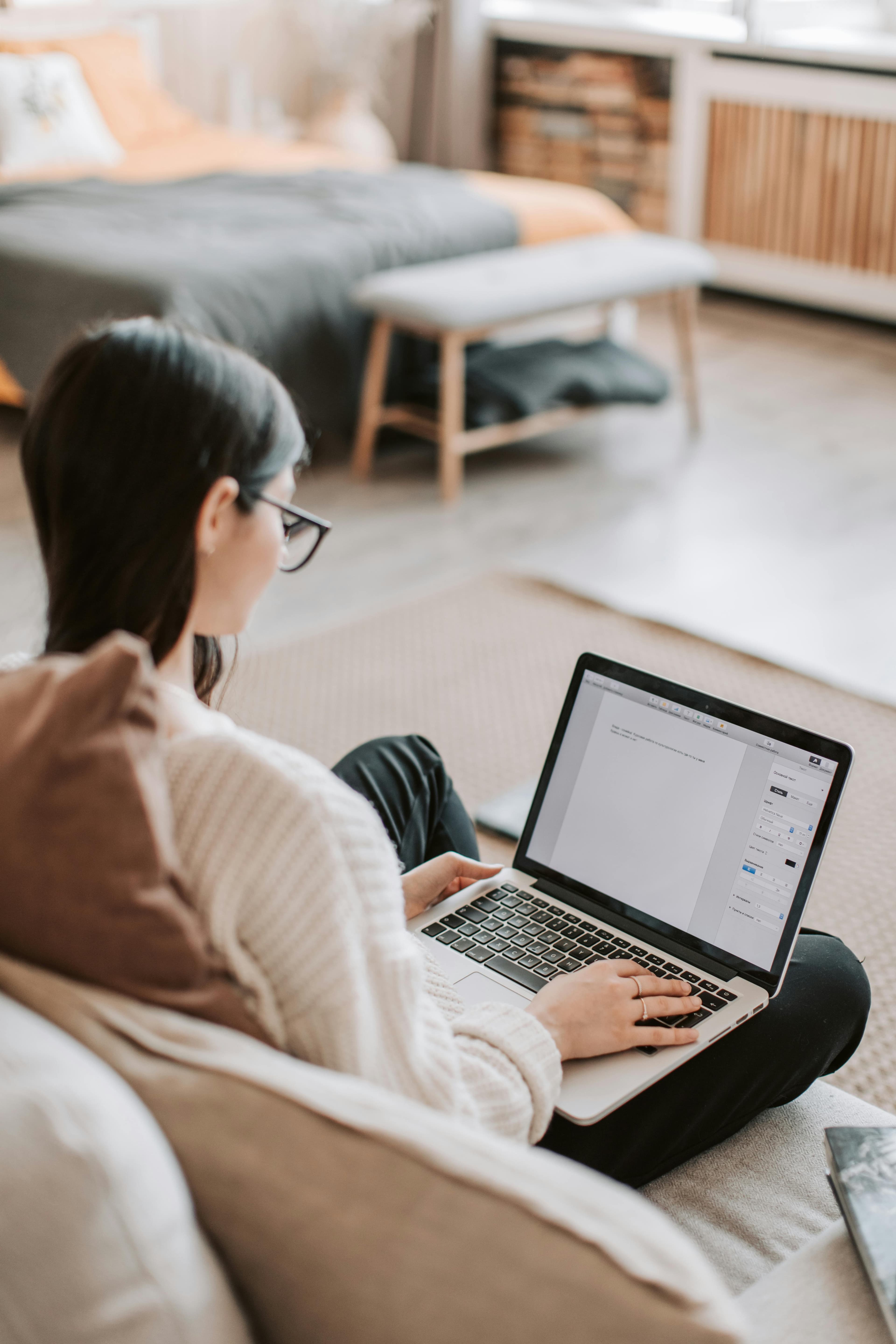 A person sitting on a sofa, typing on a laptop in a cozy living room. The individual is wearing glasses and appears to be working on content or software. This environment suggests a focus on productivity, possibly utilizing A/B testing software for digital projects.