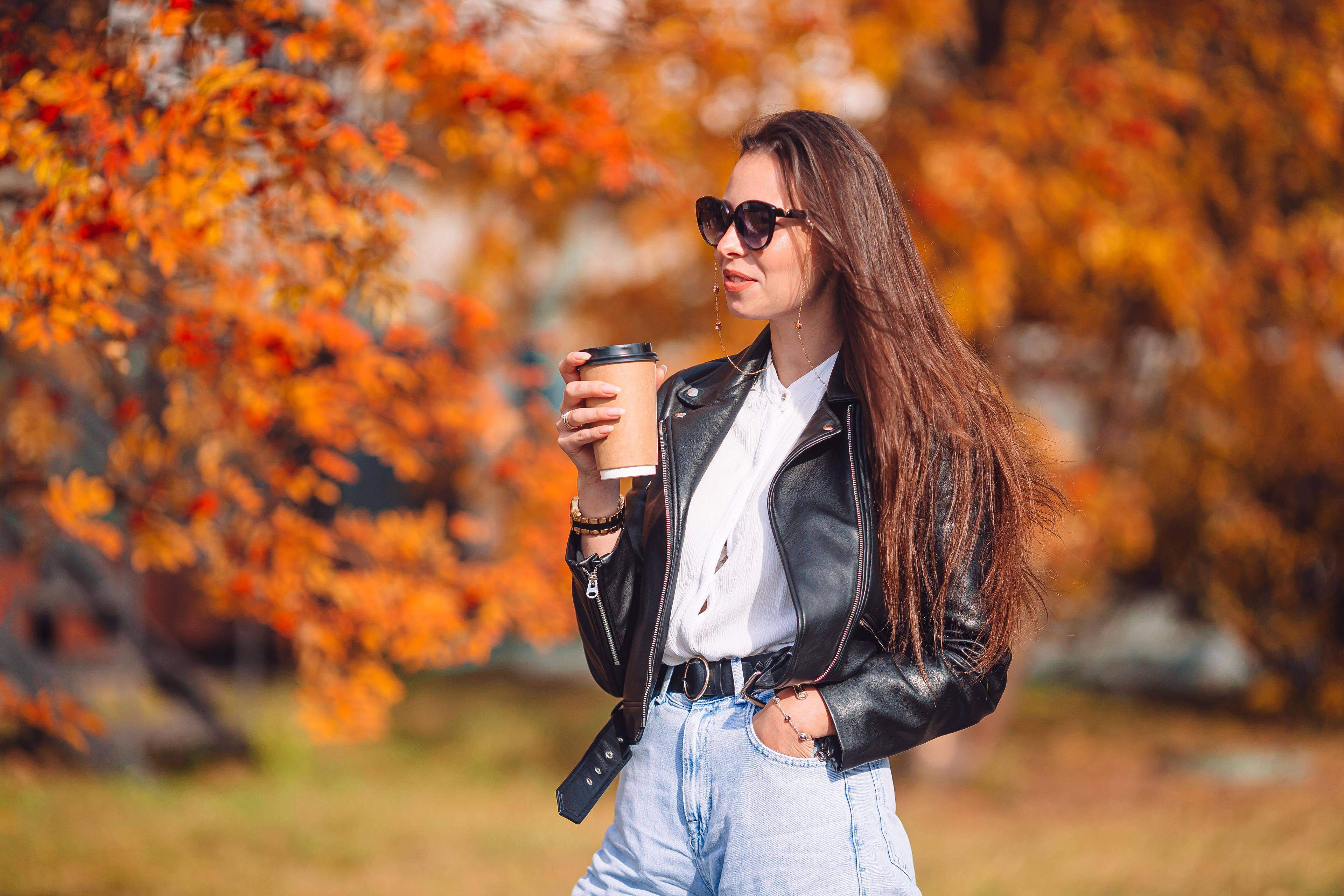 A woman wearing a black leather jacket, white blouse, and sunglasses holds a coffee cup while standing outside in front of vibrant autumn foliage. The scene conveys a sense of relaxed, stylish enjoyment of the fall season. This image represents one dataset in the concept of "sklearn train test split."