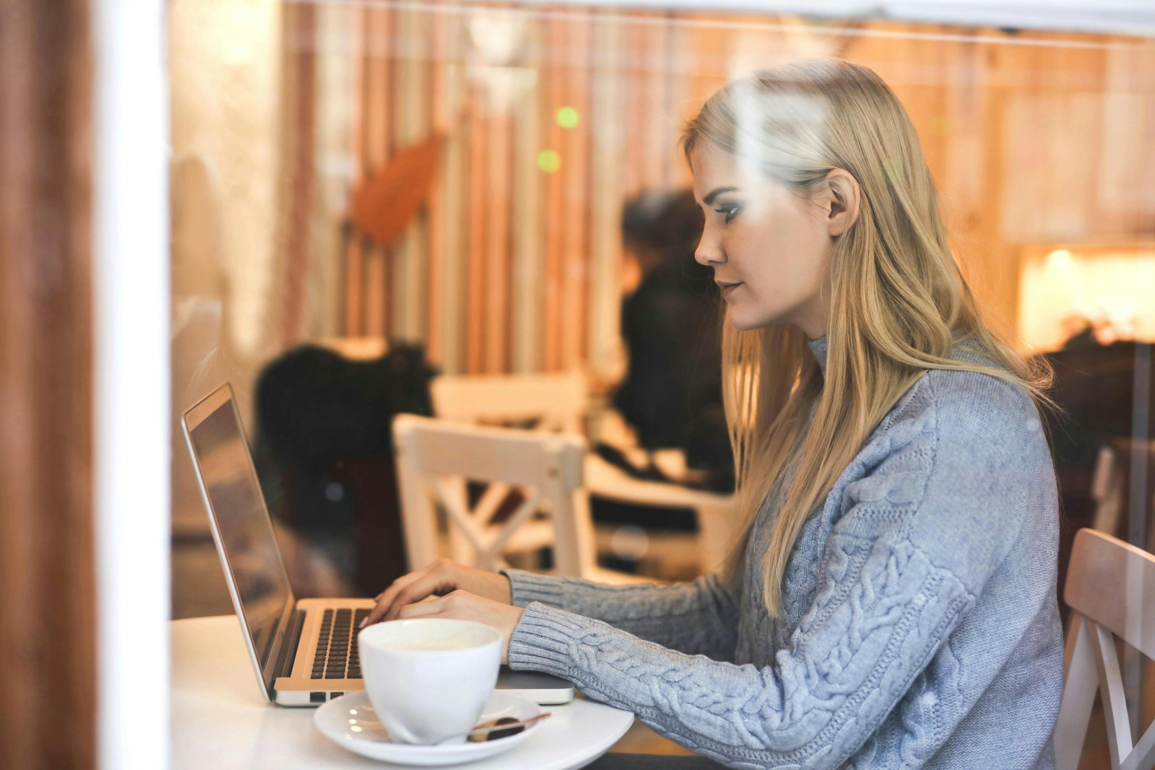 A woman sitting at a café table with a laptop, working intently with a cup of coffee beside her, illustrating the work environment for influencer marketing agencies.