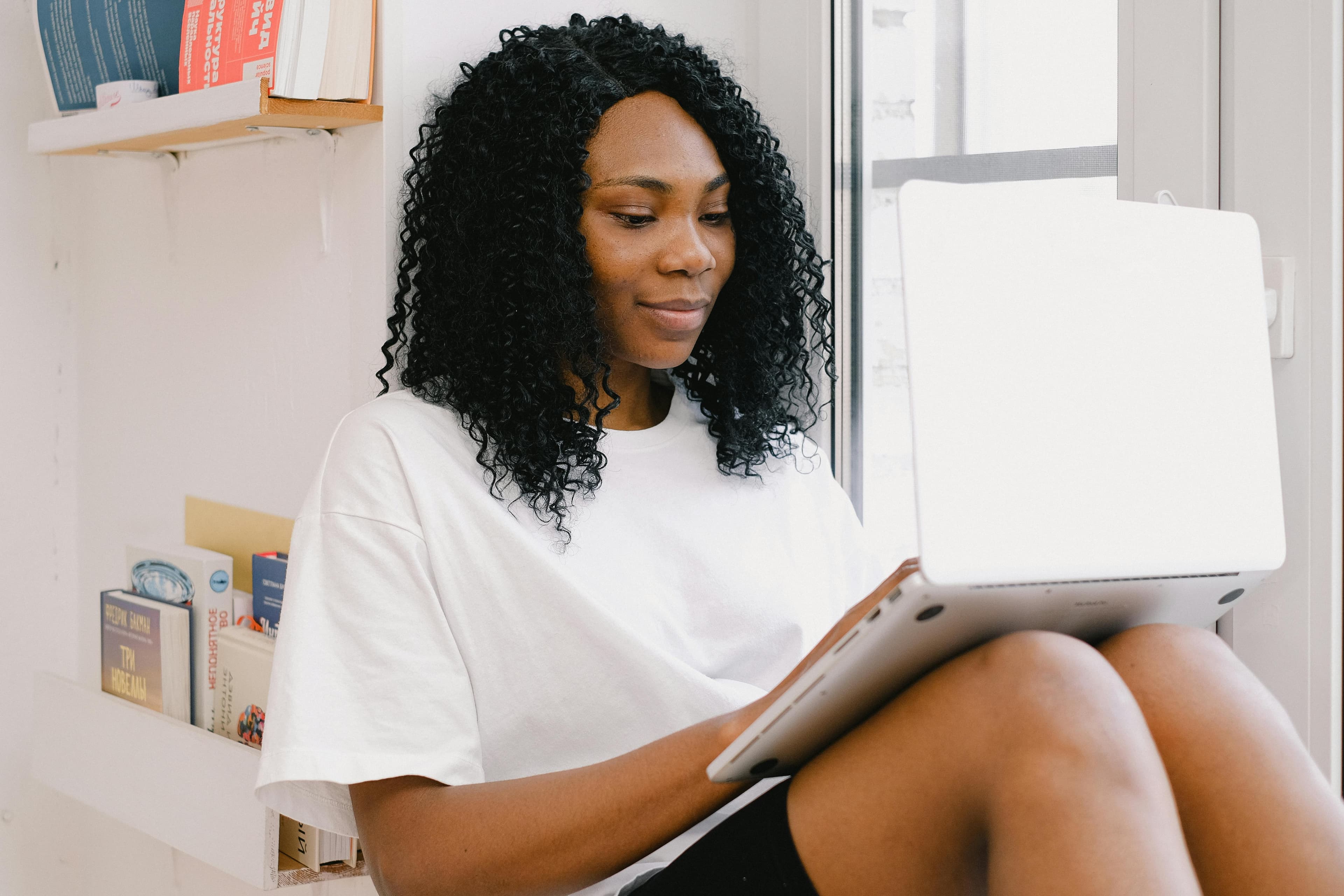 A woman with curly hair wearing a white t-shirt, sitting by a window and working on a laptop. She appears focused and engaged, likely involved in tasks related to event marketing jobs. Behind her, a bookshelf with various books adds a touch of academia and inspiration to the setting. The natural light from the window creates a bright and inviting workspace, ideal for creative and strategic planning.