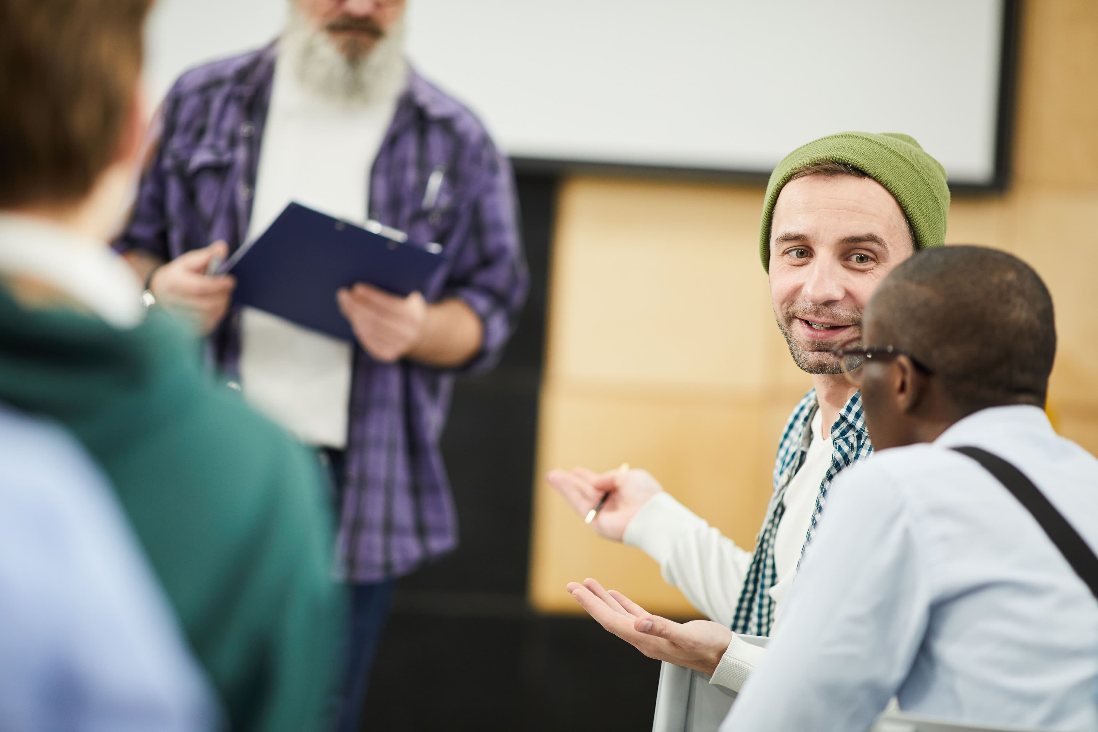 Two people in a discussion, one gesturing with a pen while another listens attentively, with a third person in the background holding a clipboard. This setting illustrates "small business marketing consultants" engaging in collaborative planning or brainstorming sessions with clients to enhance their marketing approaches.