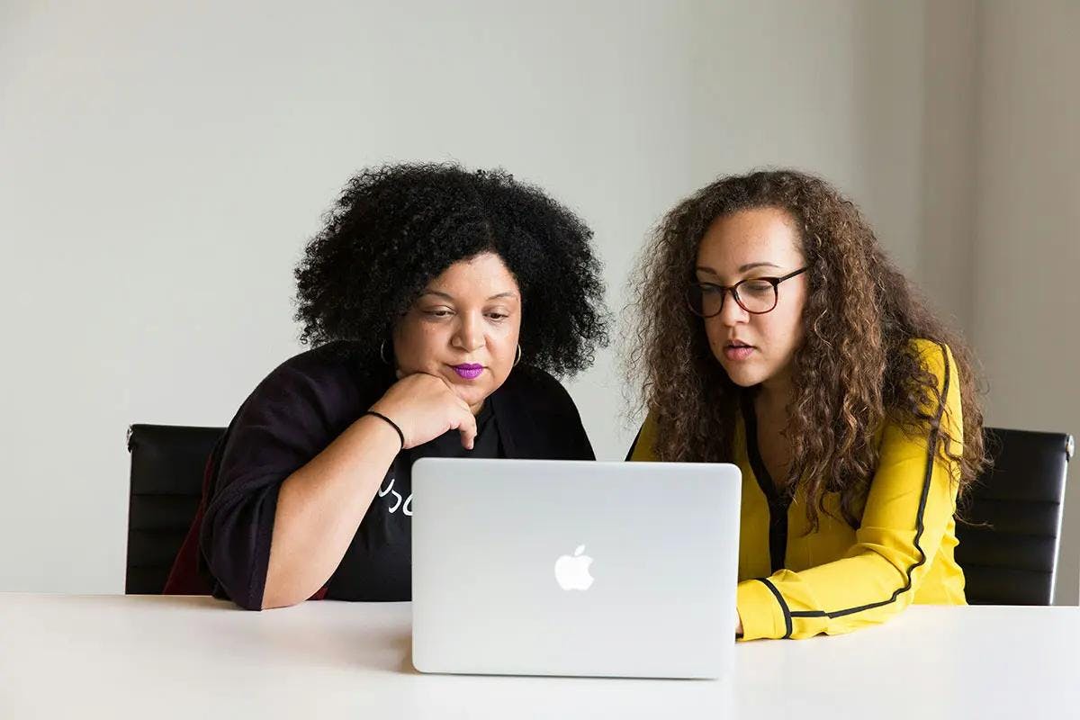 Two women are sitting at a white table, focused on a laptop screen in front of them. One woman, with curly hair and wearing a black top, is leaning on her hand thoughtfully. The other woman, with long curly hair and glasses, is wearing a yellow shirt and is engaged in the content on the screen. The background is a plain, light-colored wall, creating a professional and minimalist environment. This image is suitable for illustrating the concept of "Bing AI image generator."
