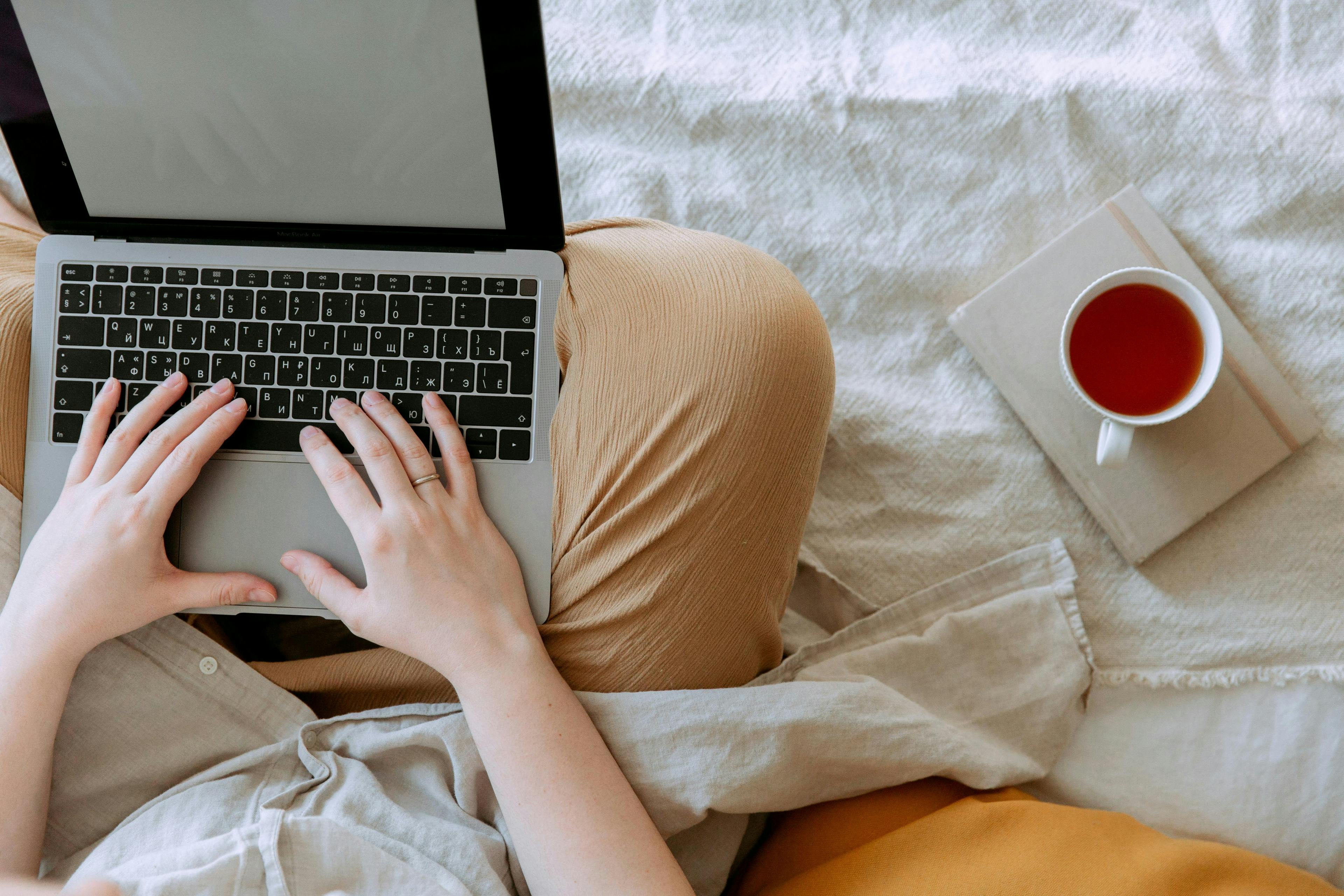 a person sitting on a bed, typing on a laptop, with a cup of tea placed on a closed book beside them. This serene setting prompts the question: "Is lead magnetic?"