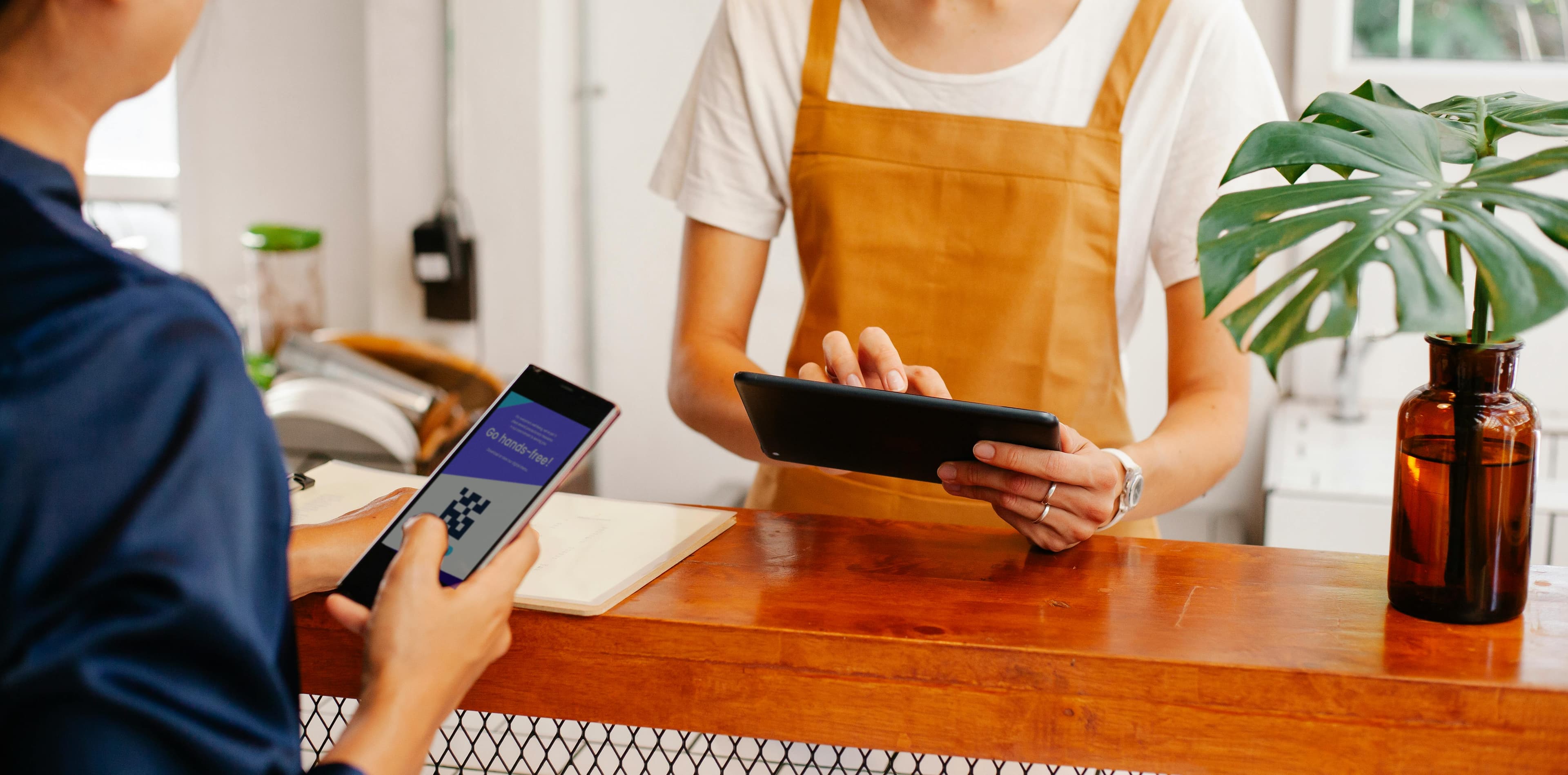 A store clerk in a tan apron uses a tablet while a customer holds up a smartphone displaying a QR code for hands-free payment. Ideal for illustrating seamless customer experiences enhanced by an AI landing page builder, designed for mobile responsiveness and engagement.