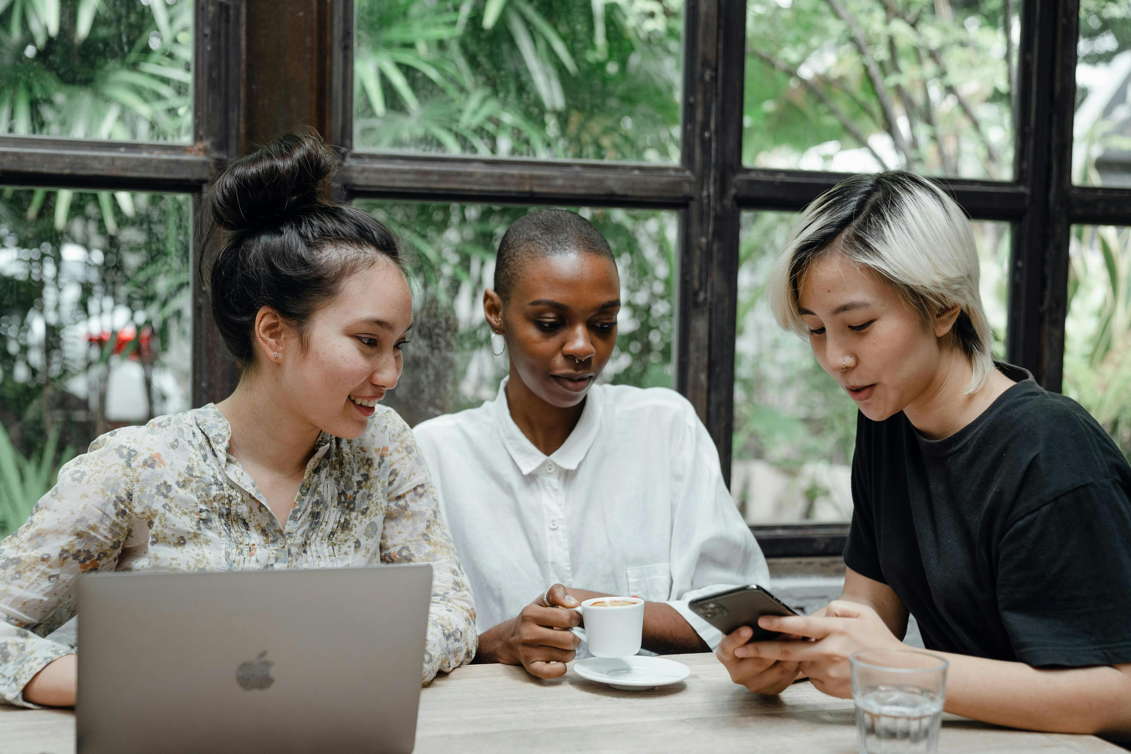Three diverse women are seated at a table, engaging in a collaborative discussion. One has a laptop open, another holds a coffee cup, and the third is showing something on her phone. The setting is bright with a natural backdrop, ideal for illustrating the concept of "influencer marketing platforms."