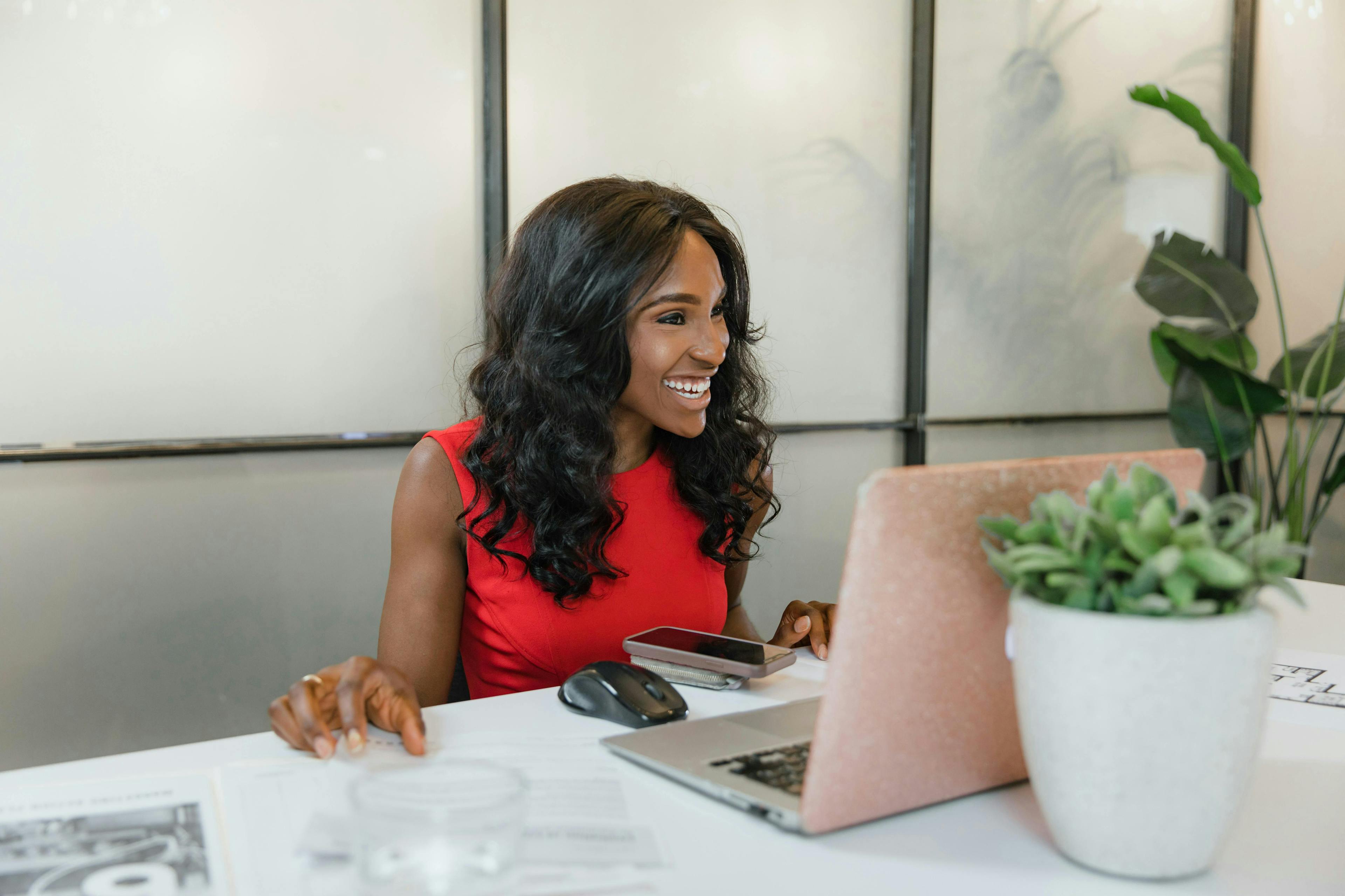 A smiling woman in a red dress works on a laptop at a desk, with a smartphone and documents nearby. A small potted plant is on the desk. Digital marketing agency for startups.