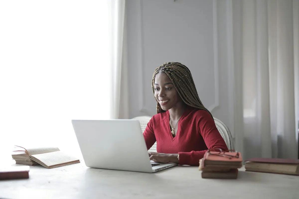 A woman with long braided hair sits at a well-lit desk, smiling as she works on her laptop. She is wearing a red sweater and appears engaged and focused. On the desk are several open books and notebooks, suggesting a productive work session. The setting is bright and professional, making this image suitable for illustrating the concept of a "business marketing coach."