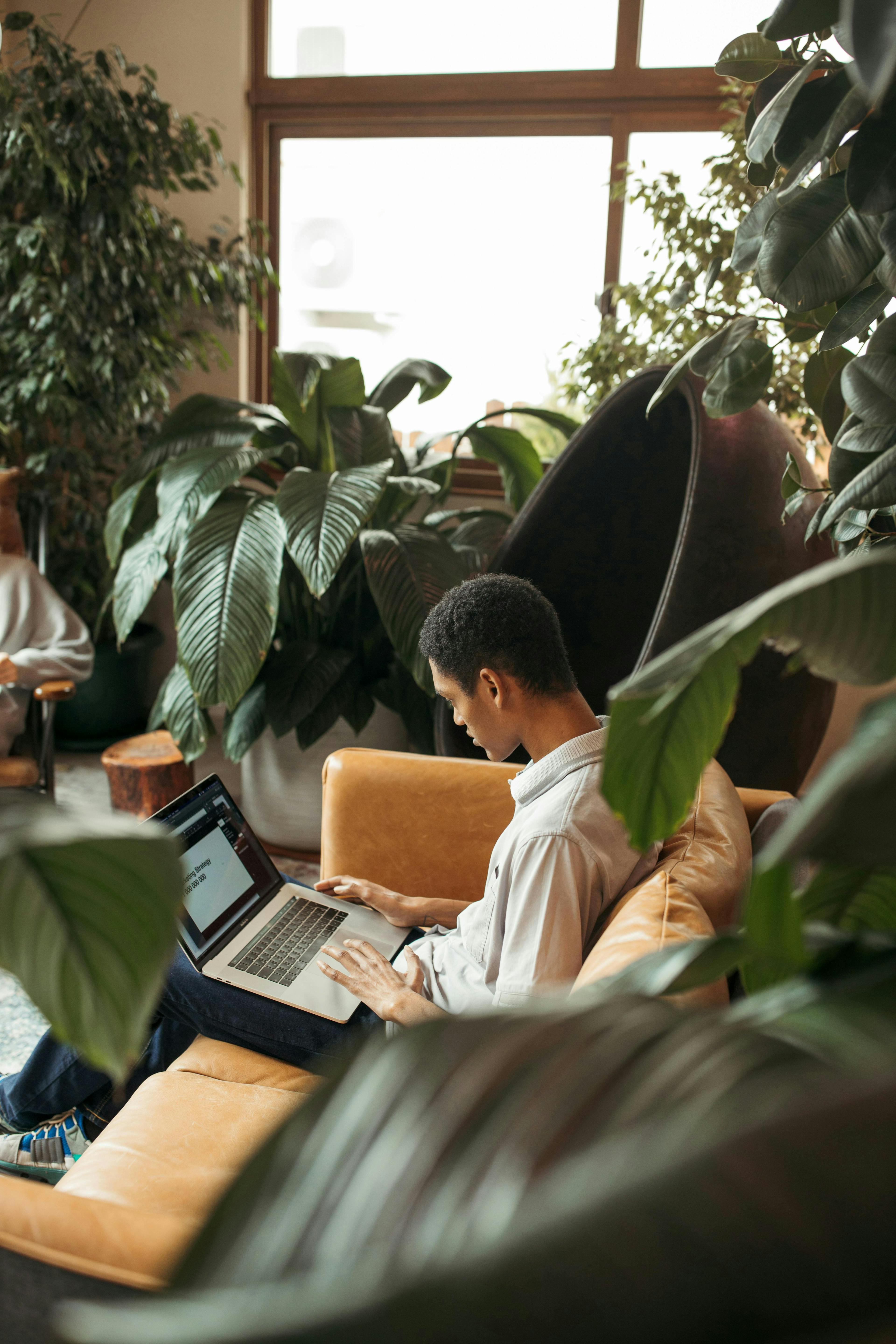 a person sitting on a comfortable couch surrounded by lush indoor plants, working on a laptop. This relaxed and inviting space is suitable for exploring various home improvement products, including a "lawn mower track conversion kit."